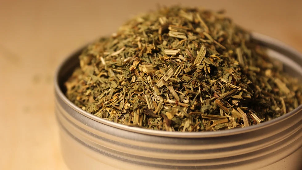 Close-up of organic California poppy loose-leaf tea by Healing Herbals, showing a textured blend of dried green leaves and stems piled in a clear round container on a warm neutral background.