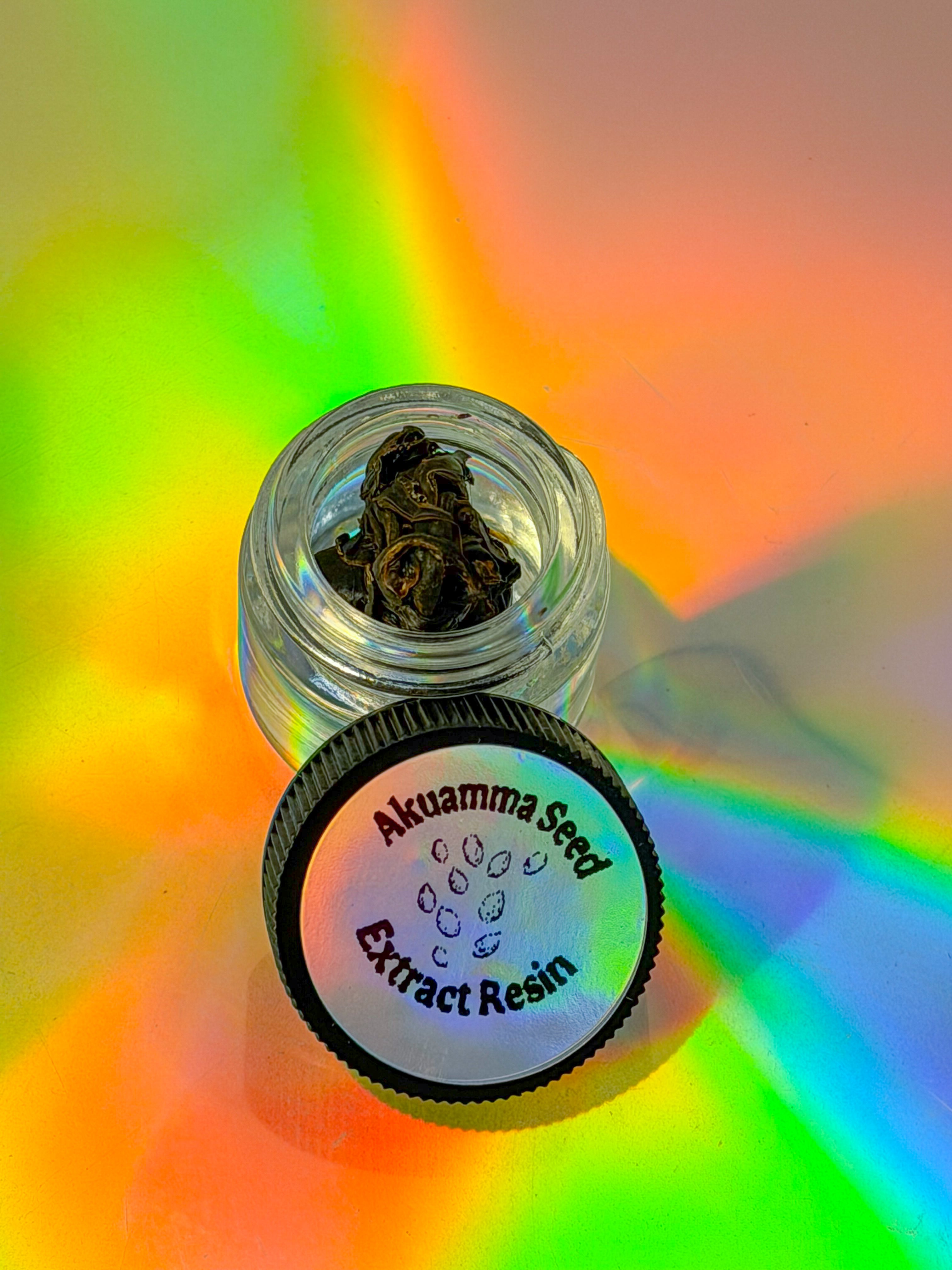 Jar of Akuamma Seed Extract Resin on a colorful background. The resin is a brown, stringy resin in a clear jar, with a black lid next to it labeled "akuamma seed extract resin"