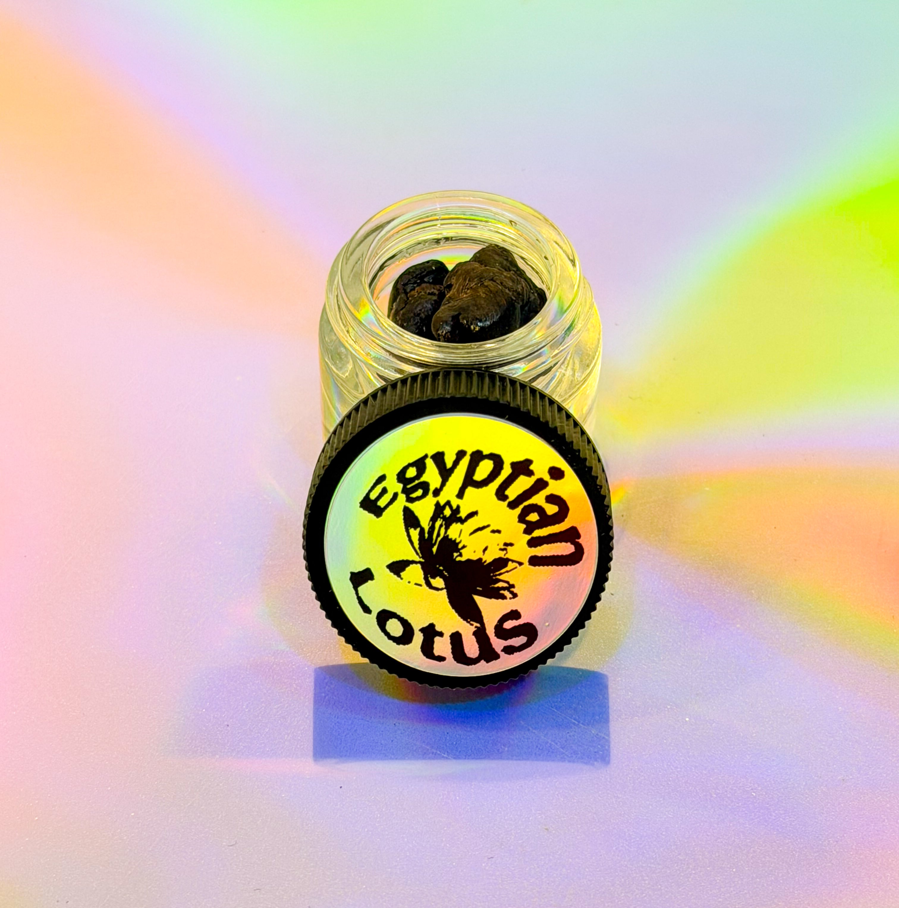 Center frame is a clear jar with brown resin from the egyptian blue lotus plant. A black jar sits besides labeled "egyptian lotus" in a yellow reflective gradient. The jar sits on a reflective colorful gradient.