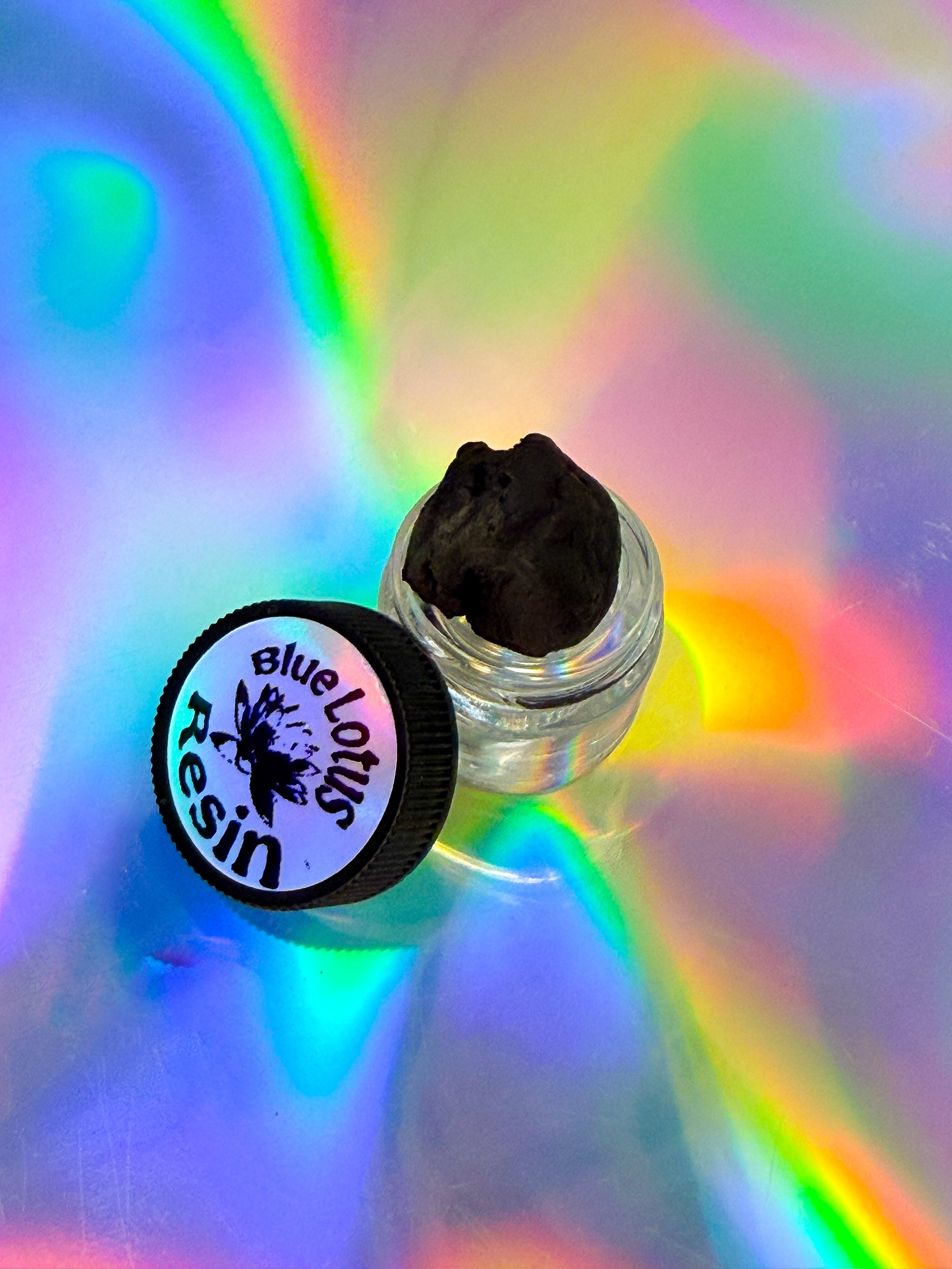 Small clear glass jar containing a dark resin piece, placed beside a black lid labeled “Blue Lotus Resin,” set on an iridescent background with vibrant rainbow reflections.