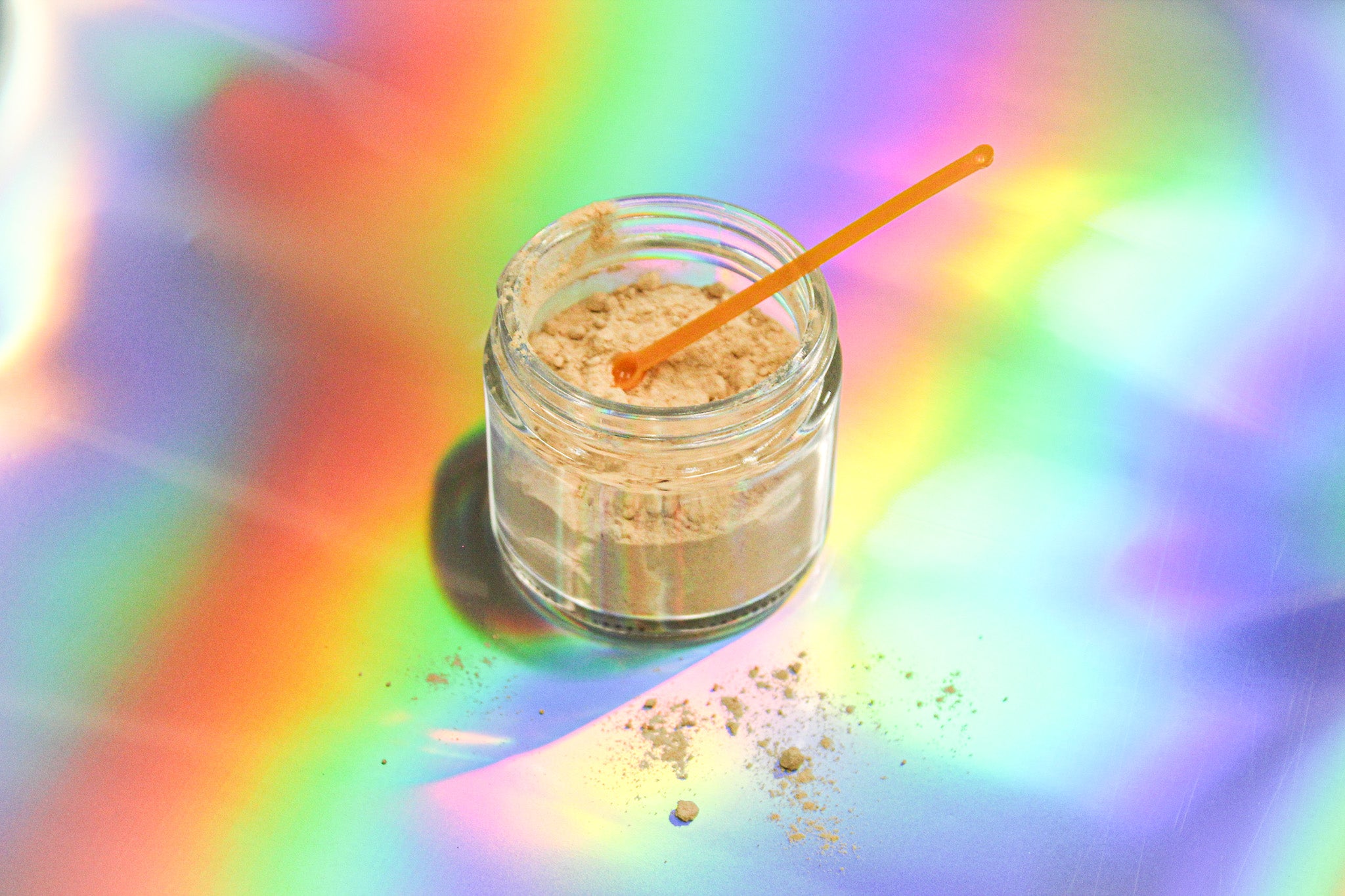 IX kanna extract powder by Healing Herbals in a small clear glass jar, with a yellow dosing scoop resting inside and fine beige powder scattered on a vibrant iridescent rainbow background.