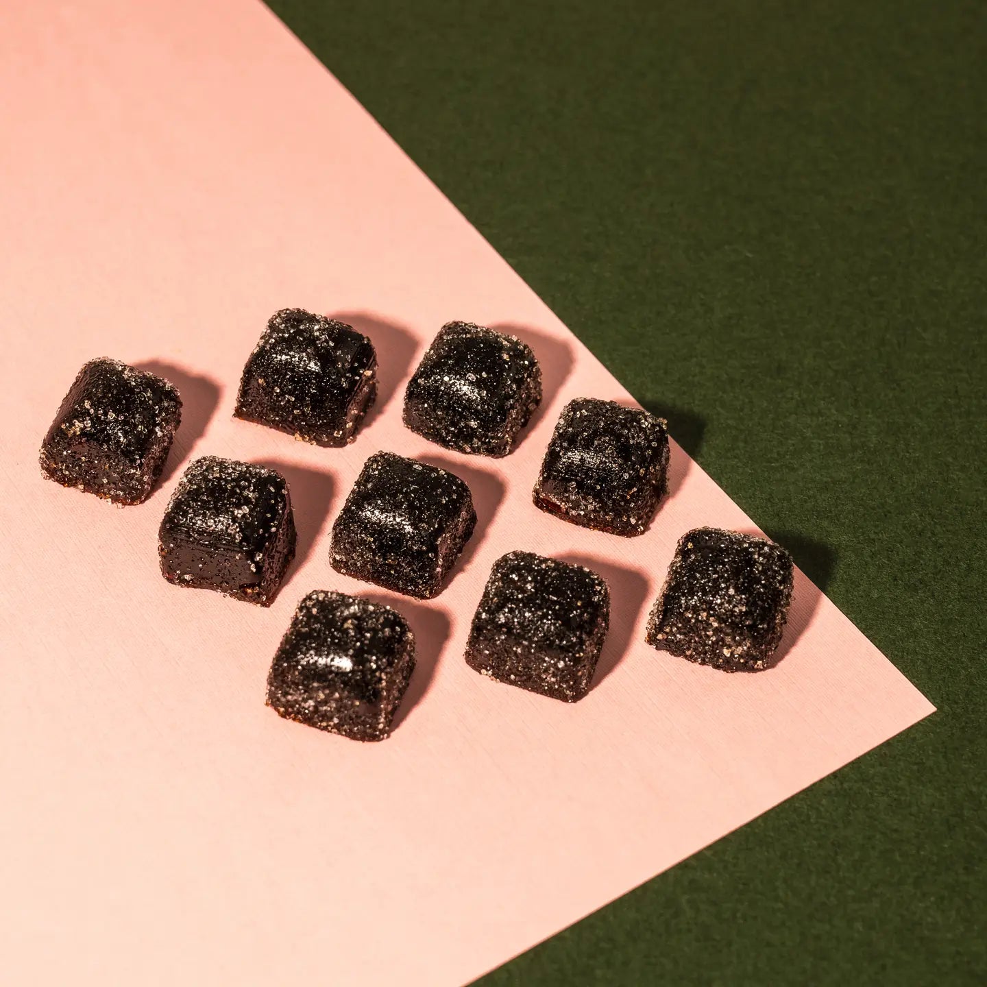 Dark square Just Relax functional gummy chews with a lightly sugared texture, arranged in a neat grid on a soft pink surface against a deep green background, photographed from above.