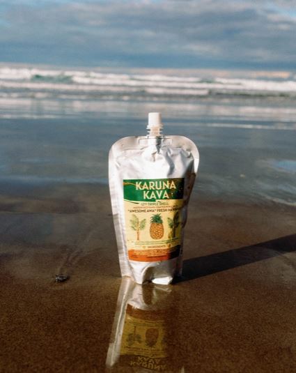 Karuna Kava Fresh Green Hawaiian Awa in a silver spouted pouch standing on wet beach sand, with ocean waves and cloudy sky in the background, reflected on the shoreline.