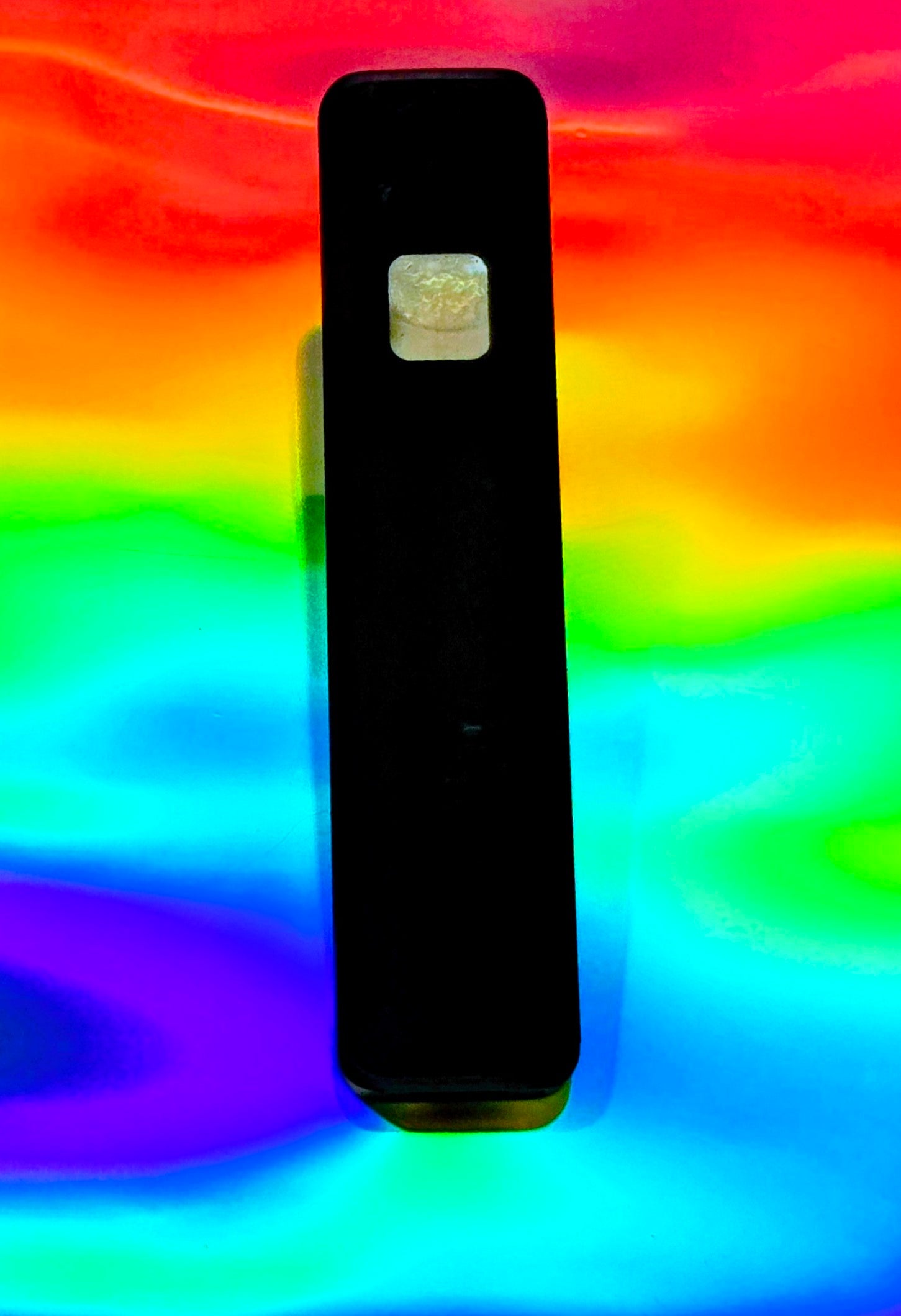 Slim black disposable vape device with a small viewing window showing light-colored herbal extract, standing upright against a vivid rainbow holographic background.