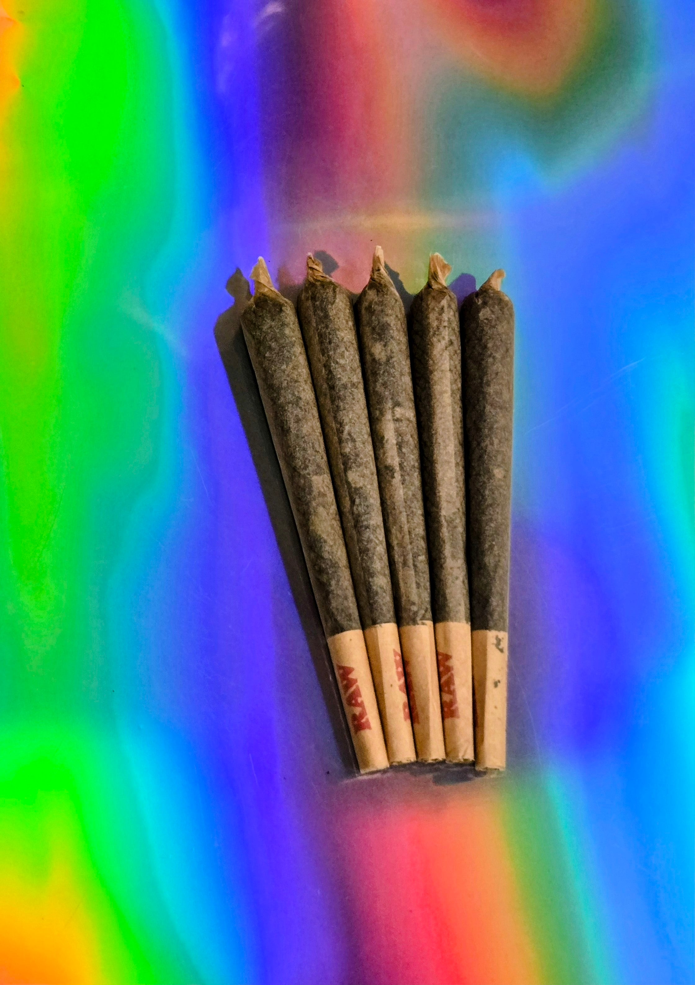 Five RAW-style pre-rolled herbal cones filled with dark botanical blend, neatly fanned on a vibrant rainbow holographic background.