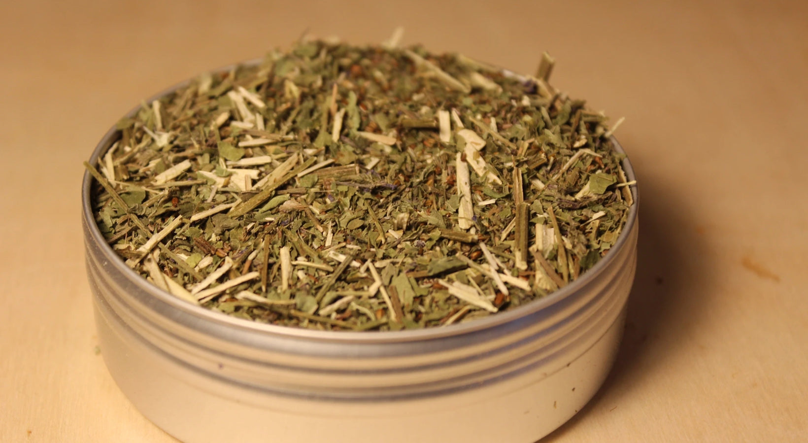 Organic blue vervain loose-leaf tea by Healing Herbals, featuring a finely cut blend of dried green leaves and pale stems packed in a clear round container on a warm wooden background.