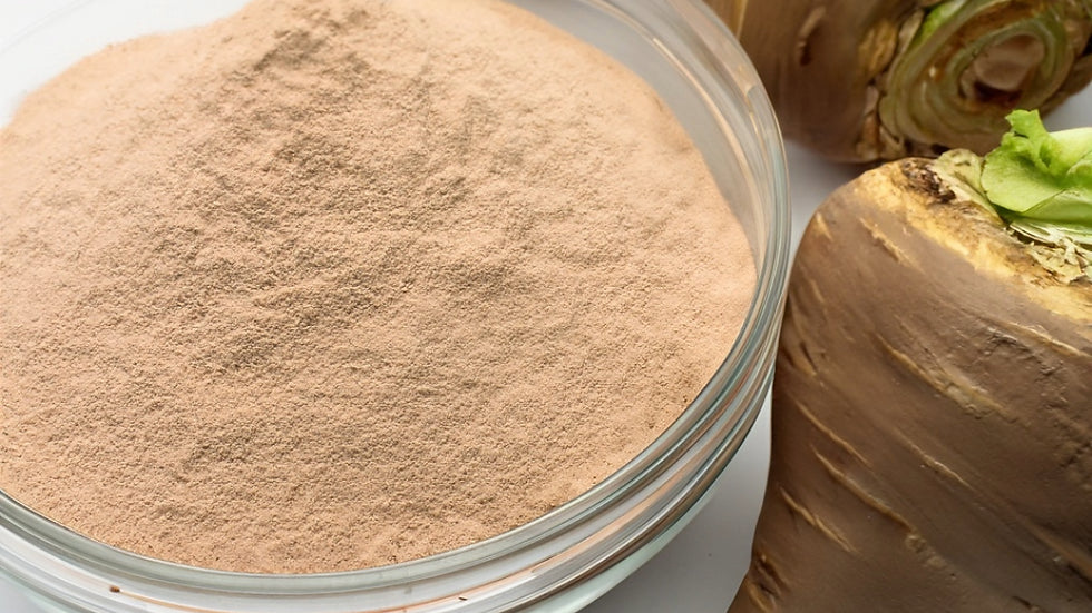 Close-up of organic maca root powder in a clear glass bowl, light tan and finely ground, with whole maca roots visible alongside on a clean white background.
