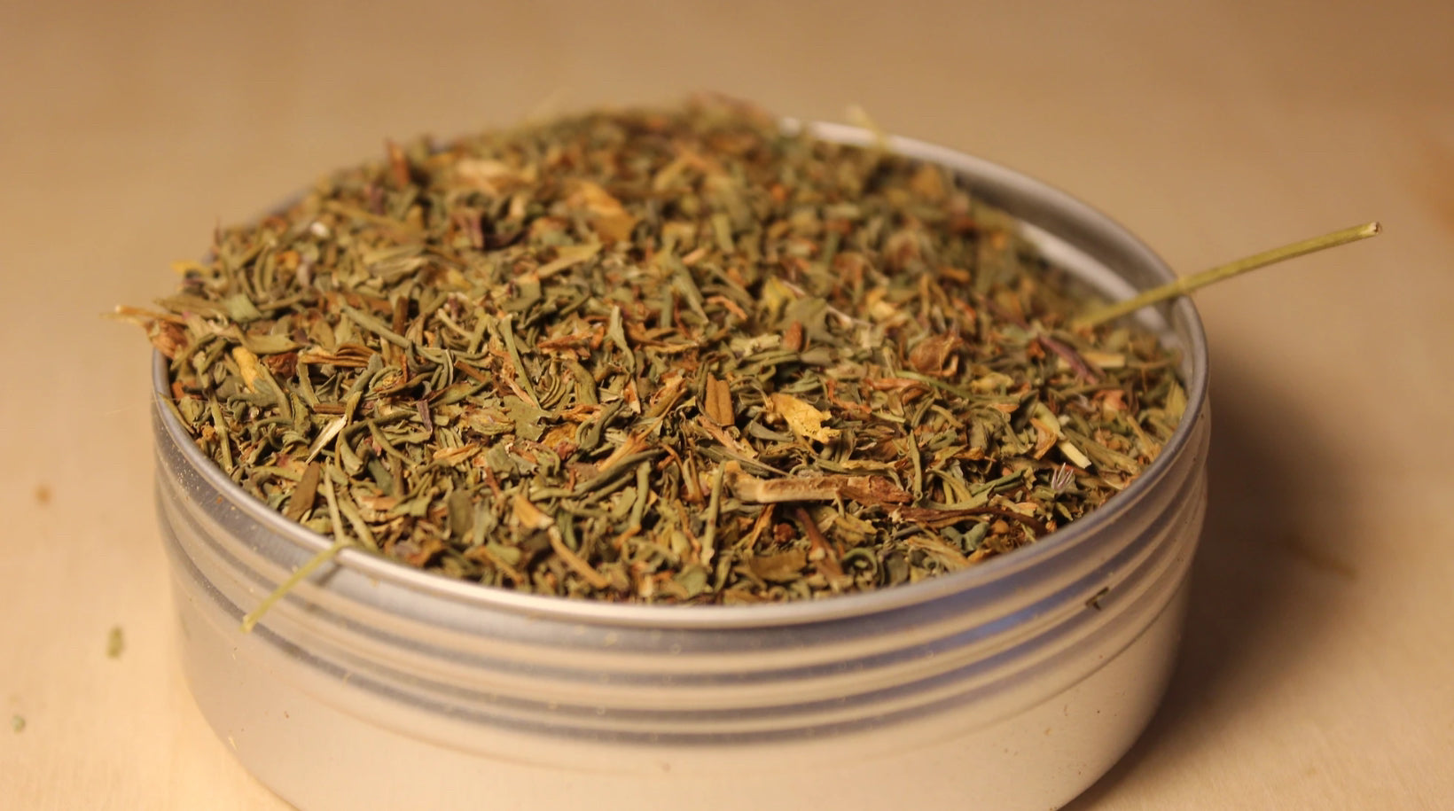 Organic St. John’s wort loose-leaf tea by Healing Herbals, showing a detailed mix of dried green leaves, stems, and small floral pieces in a clear round container on a warm neutral background.