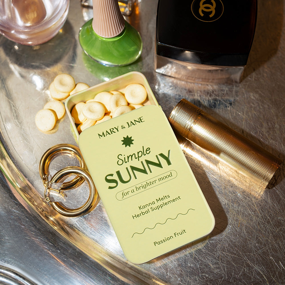 Simple Sunny Kanna Melts herbal supplement by Mary & Jane in a pale yellow pocket tin labeled “for a brighter mood,” passion fruit flavor, with round dissolvable tablets visible, styled on a metallic surface alongside personal accessories.