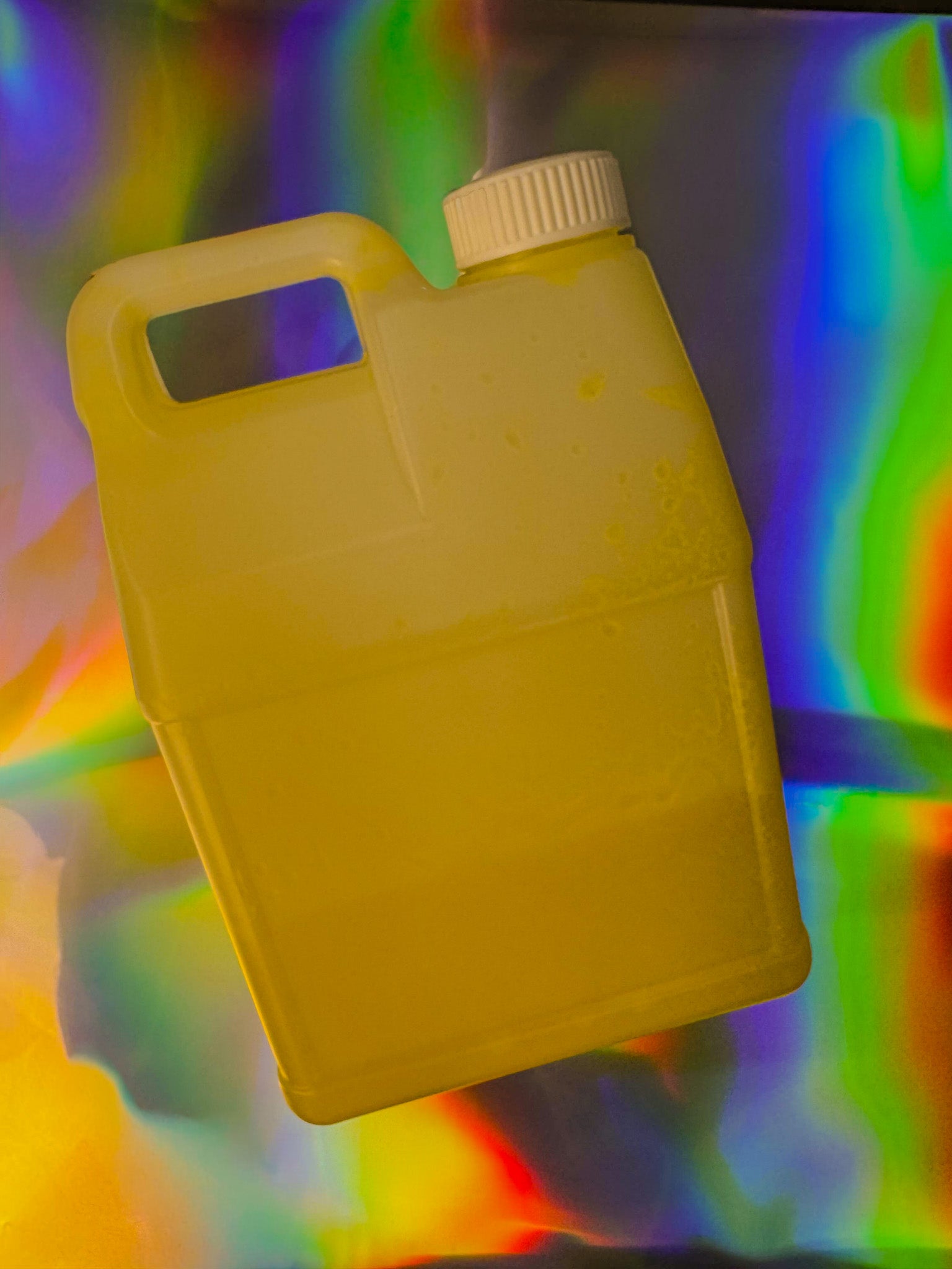 Translucent yellow liquid in a rectangular plastic jug with a built-in handle and white screw cap, photographed against a colorful iridescent rainbow background.