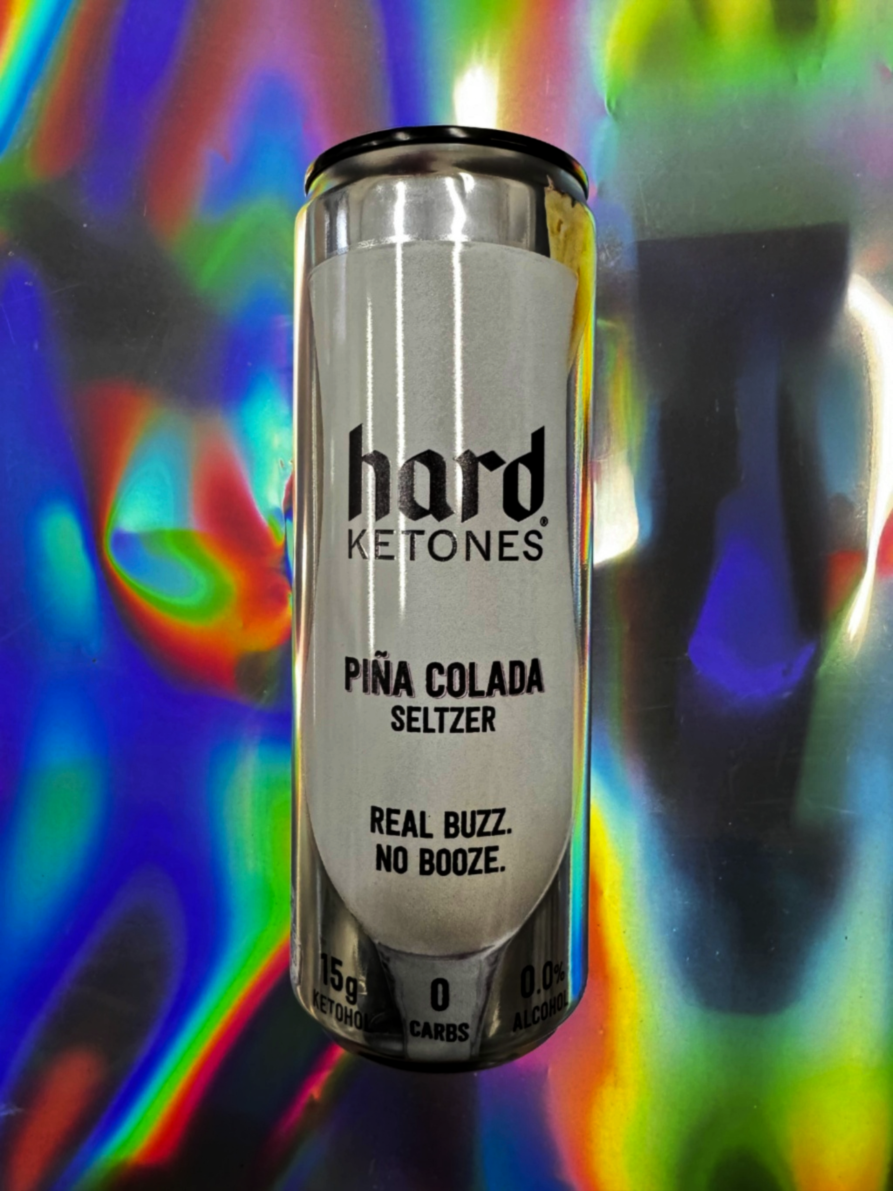 Hard Ketones Piña Colada seltzer can with silver label, featuring “Real Buzz, No Booze” text, photographed upright against a colorful iridescent rainbow background.