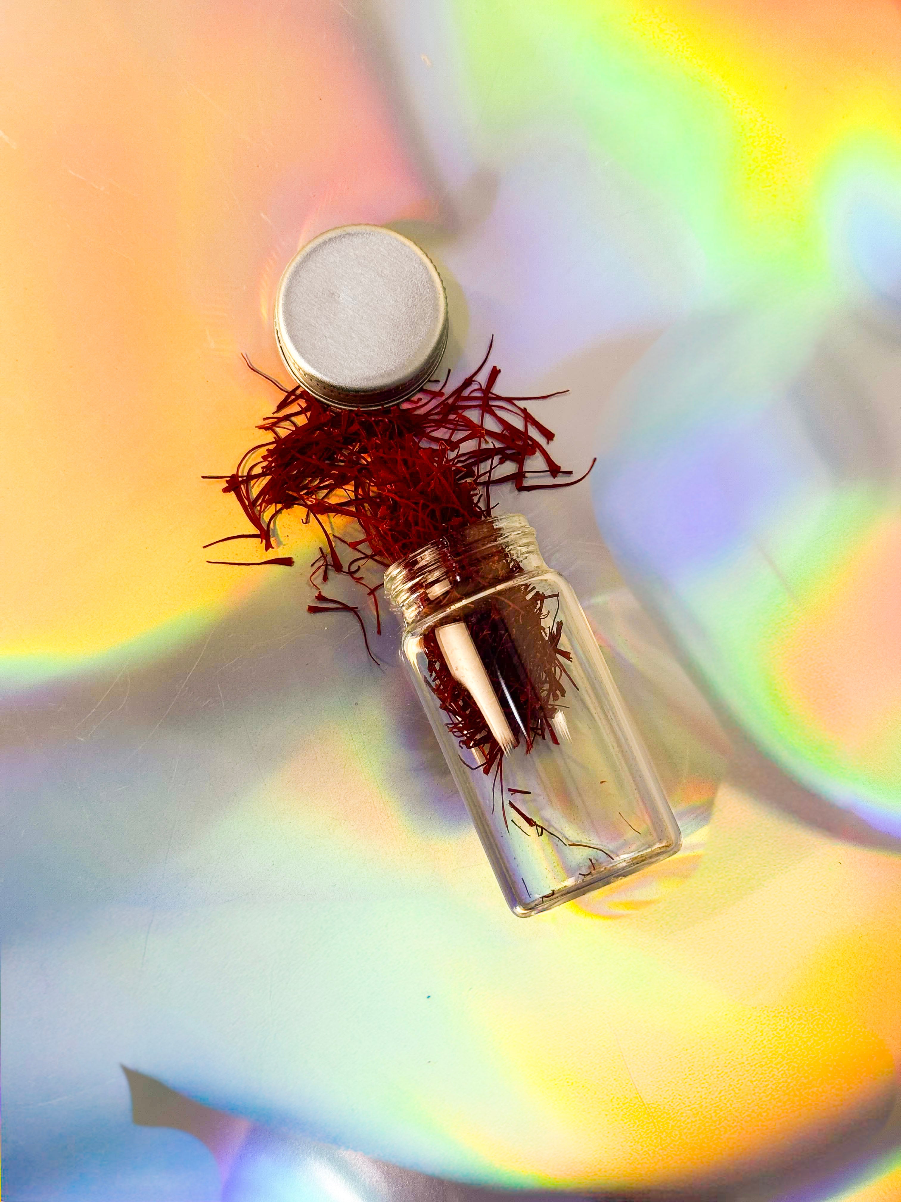 Small clear glass vial filled with deep red saffron threads, lying on its side with the silver cap removed and strands spilling out onto an iridescent rainbow background.