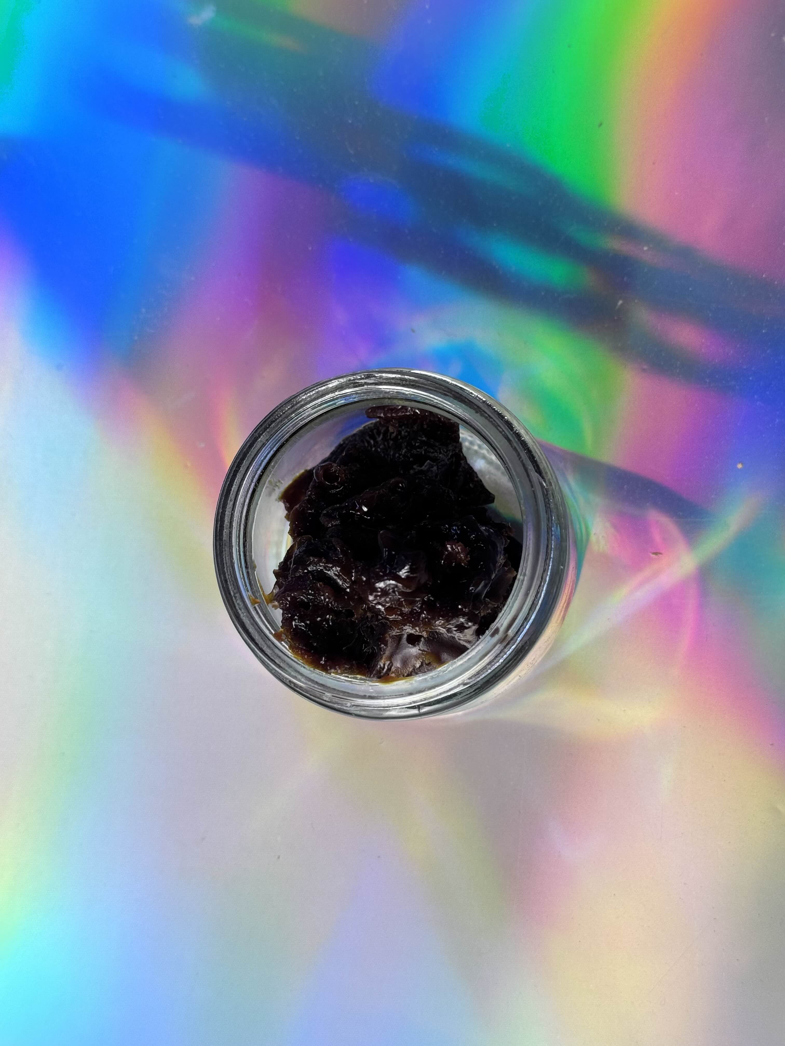 Top-down view of a clear glass jar filled with dark, glossy Amanita mushroom extract concentrate, photographed on an iridescent holographic background with rainbow light reflections.