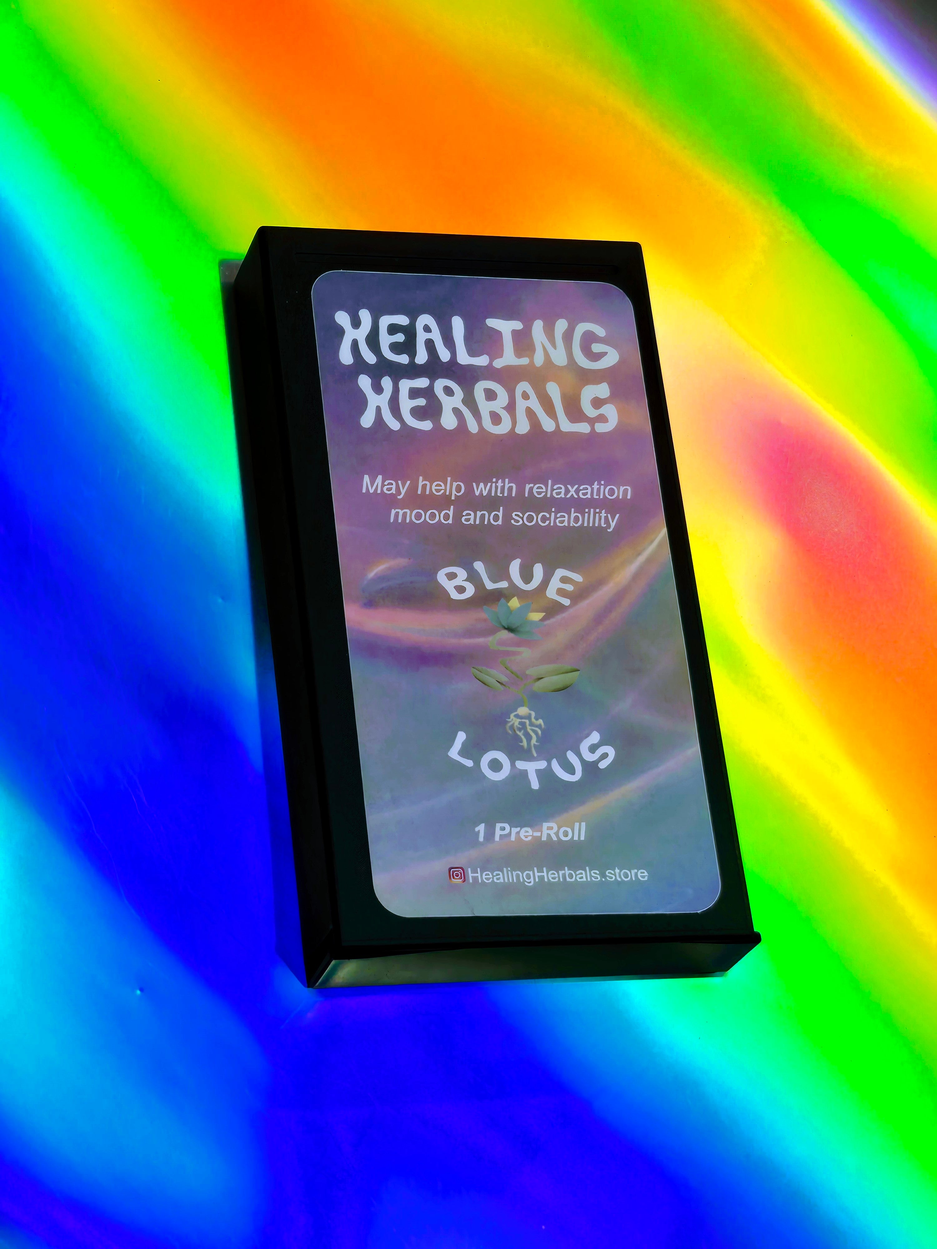 Healing Herbals Blue Lotus pre-roll box shown closed in a sleek black case with branded Blue Lotus label, photographed on a vivid iridescent rainbow background.