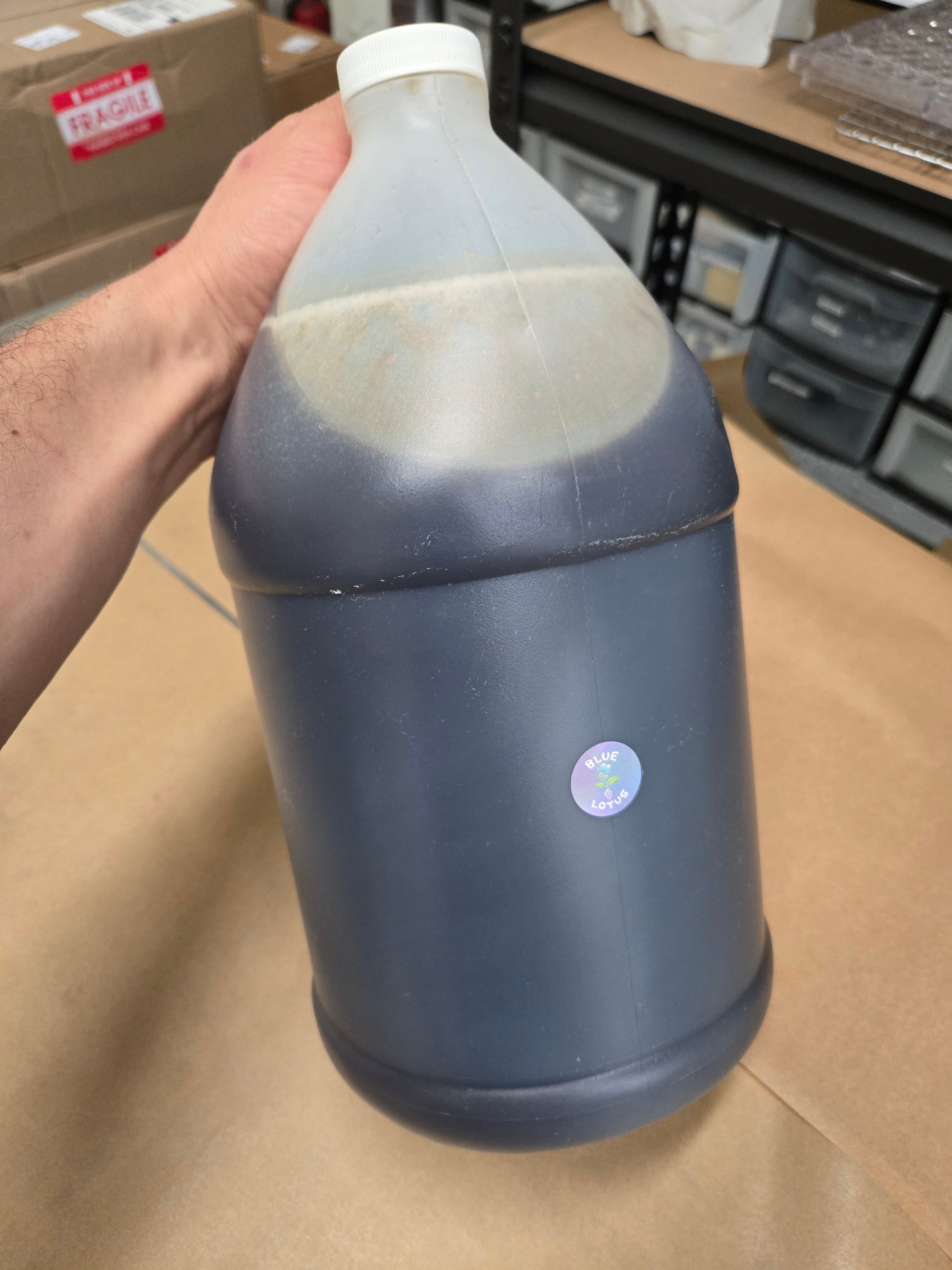 Hand holding a large translucent gallon jug filled with dark blue lotus liquid, sealed with a white cap and small round label, photographed in a warehouse setting with shelves and boxes in the background.