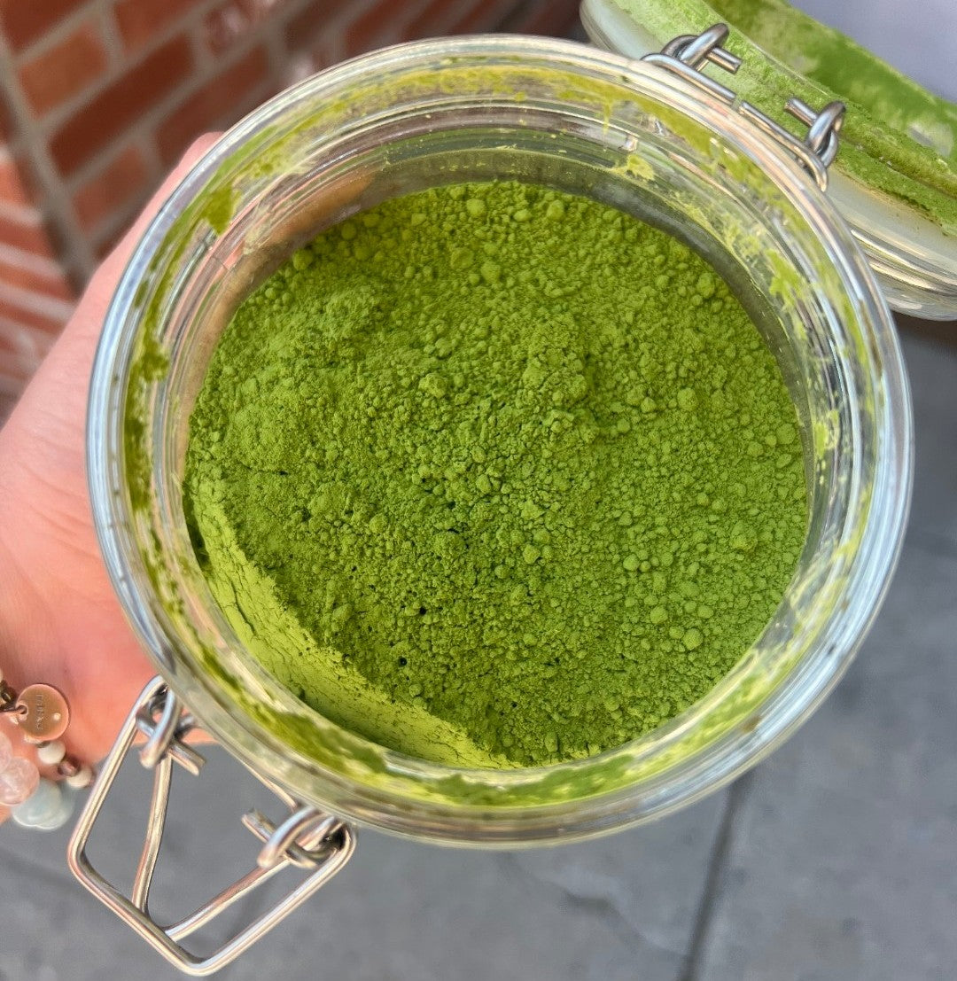 Top-down view of a clear glass jar filled with vibrant green ceremonial-grade matcha powder, showing its fine, smooth texture, with the hinged lid open and the jar held in hand against an outdoor background.