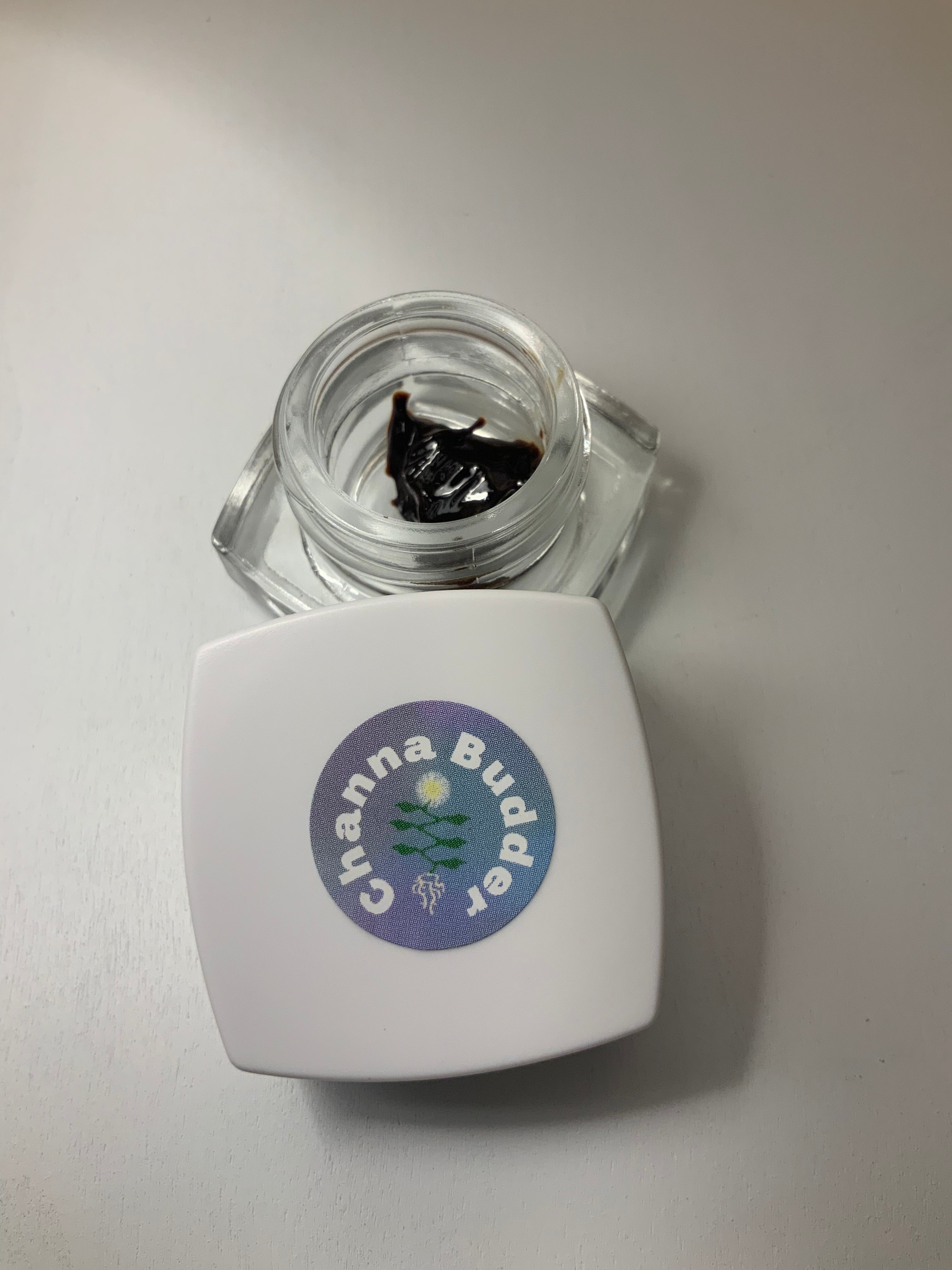Top-down view of fermented kanna extract (Channa Budder) by Healing Herbals in a clear glass jar, showing dark glossy resin with the white square lid labeled “Channa Budder” placed beside it on a clean white surface.