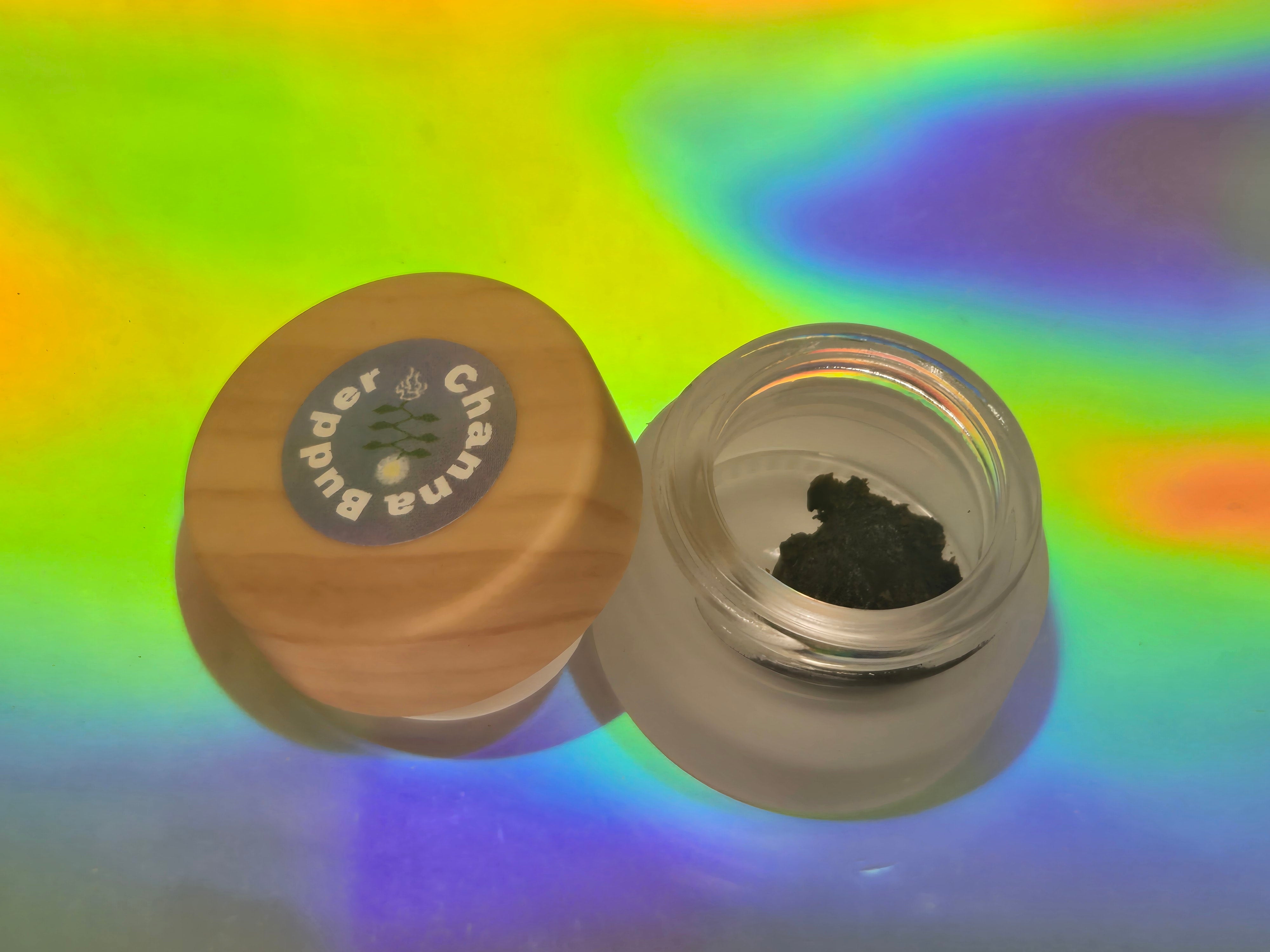 Fermented kanna extract resin (Channa Budder) by Healing Herbals in a small clear glass jar with a wooden lid, showing dark concentrated resin against a vivid iridescent rainbow background.