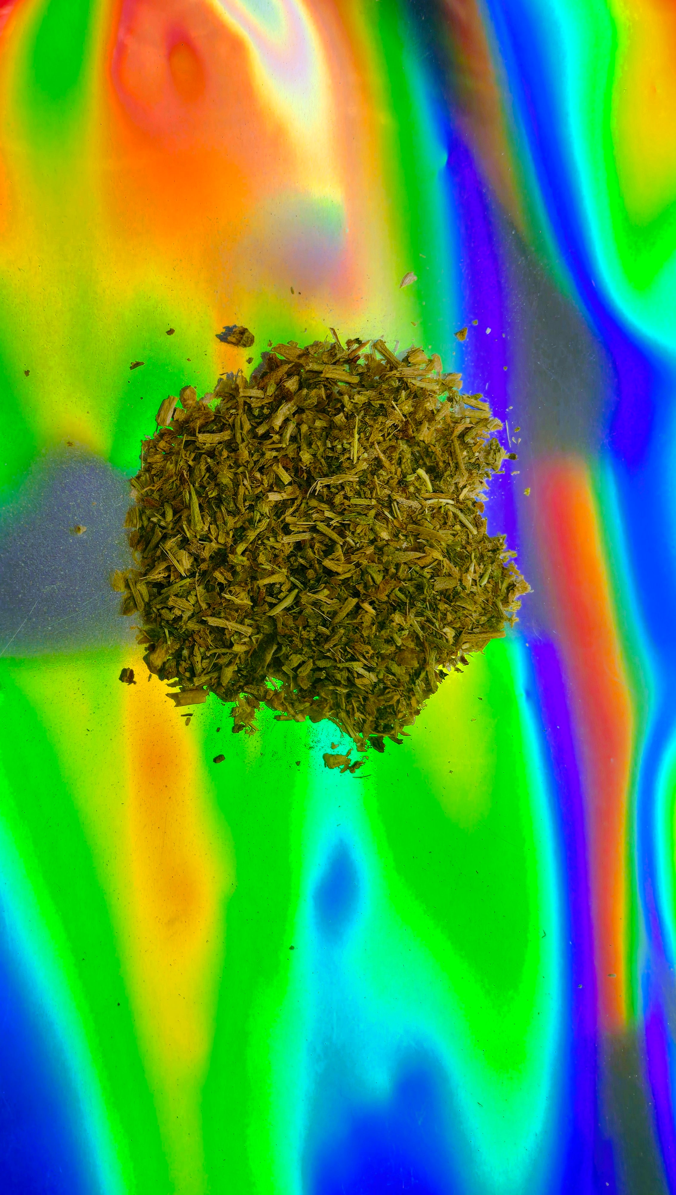 Loose-cut dried herbal leaf blend displayed in a small pile on a vibrant iridescent rainbow background, showing natural texture and plant fibers.