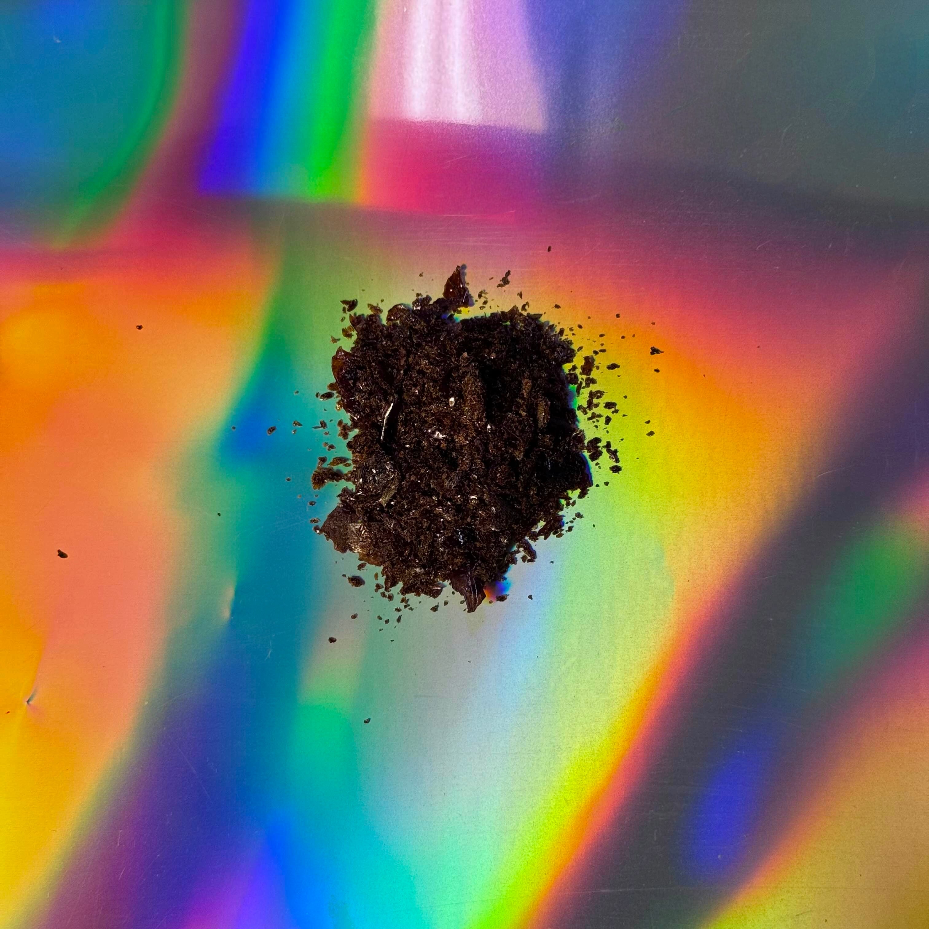 Top-down view of a small mound of dark, crumbly herbal extract placed on an iridescent holographic surface with rainbow light reflections.