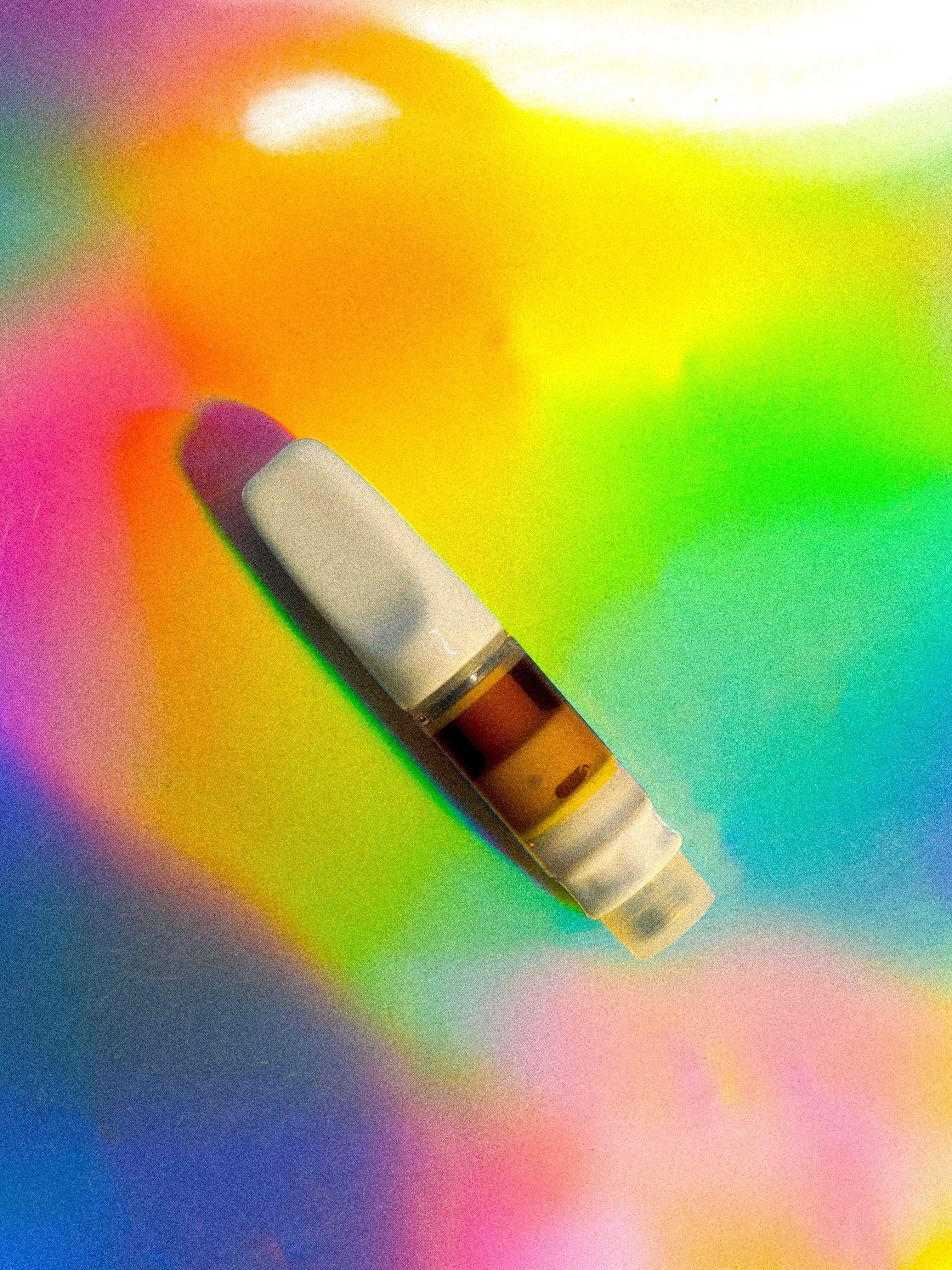 A small vape cartridge filled with amber-colored liquid, featuring a white mouthpiece and clear chamber, photographed on a vibrant iridescent rainbow background.