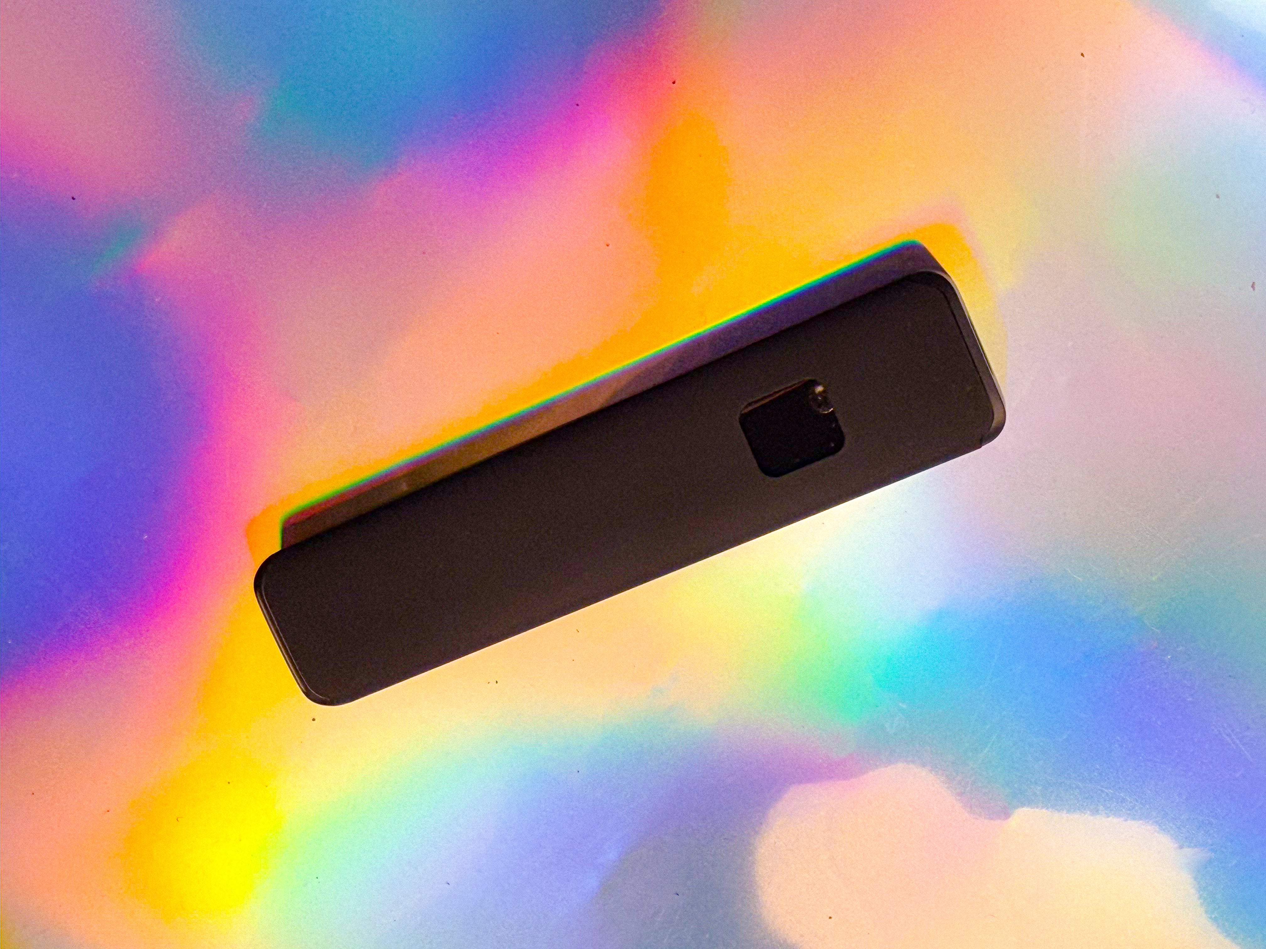 Slim black rectangular electronic device shown from above, featuring a small square detail on one side, photographed on a vibrant iridescent rainbow background.