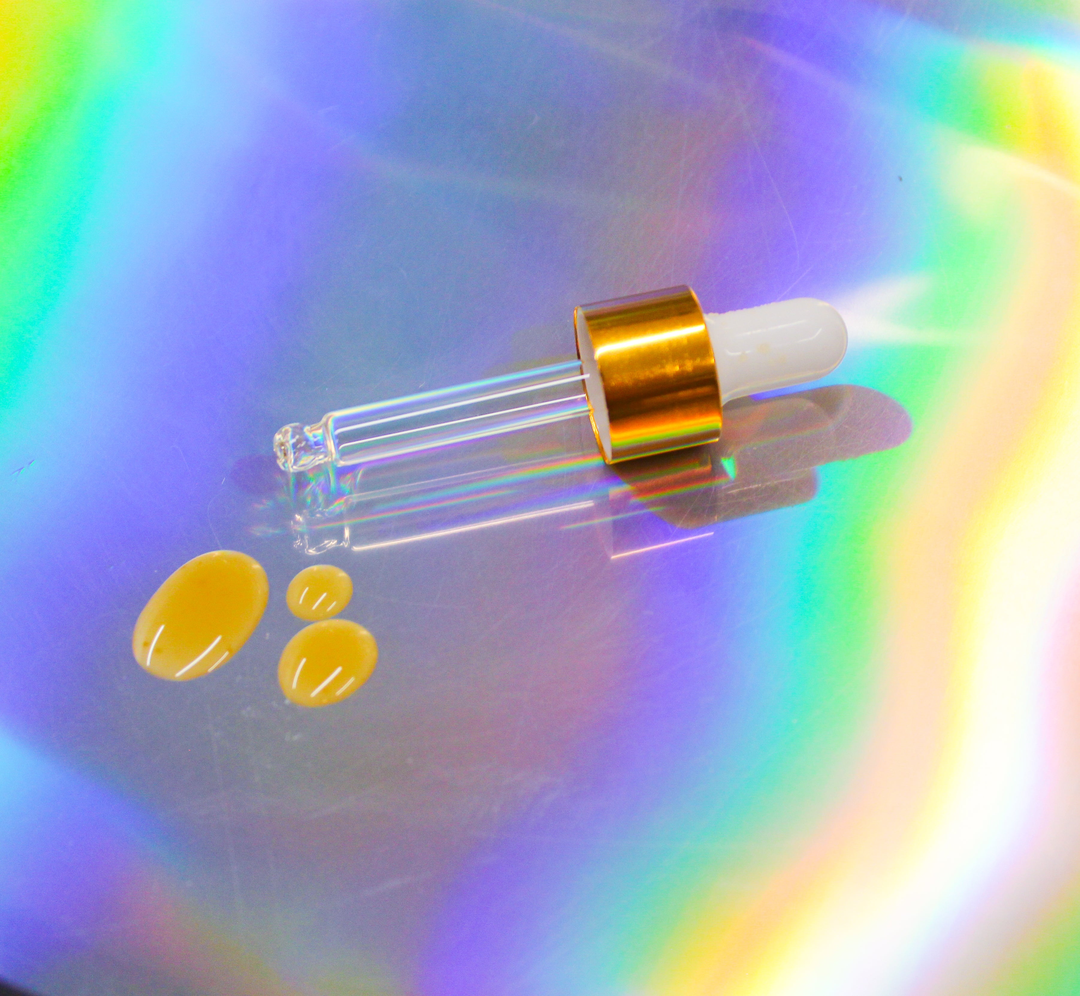 Glass dropper with gold collar and white rubber bulb resting on an iridescent holographic surface, with three glossy amber drops of Kanna liquid extract displayed beside the pipette.