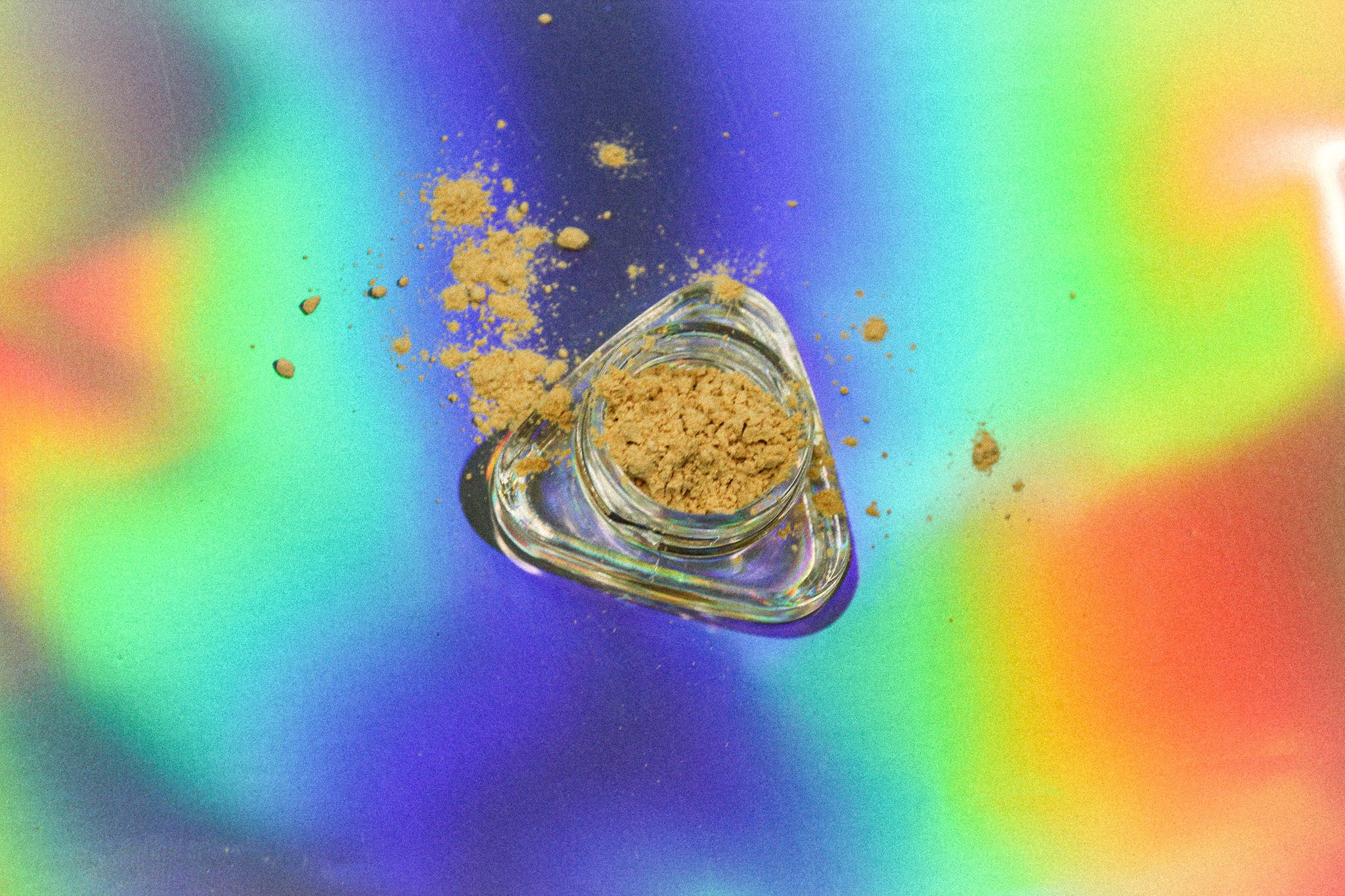 Top-down view of a small clear glass jar filled with fine kanna extract powder, with scattered tan granules around it, photographed on a vibrant iridescent rainbow background.