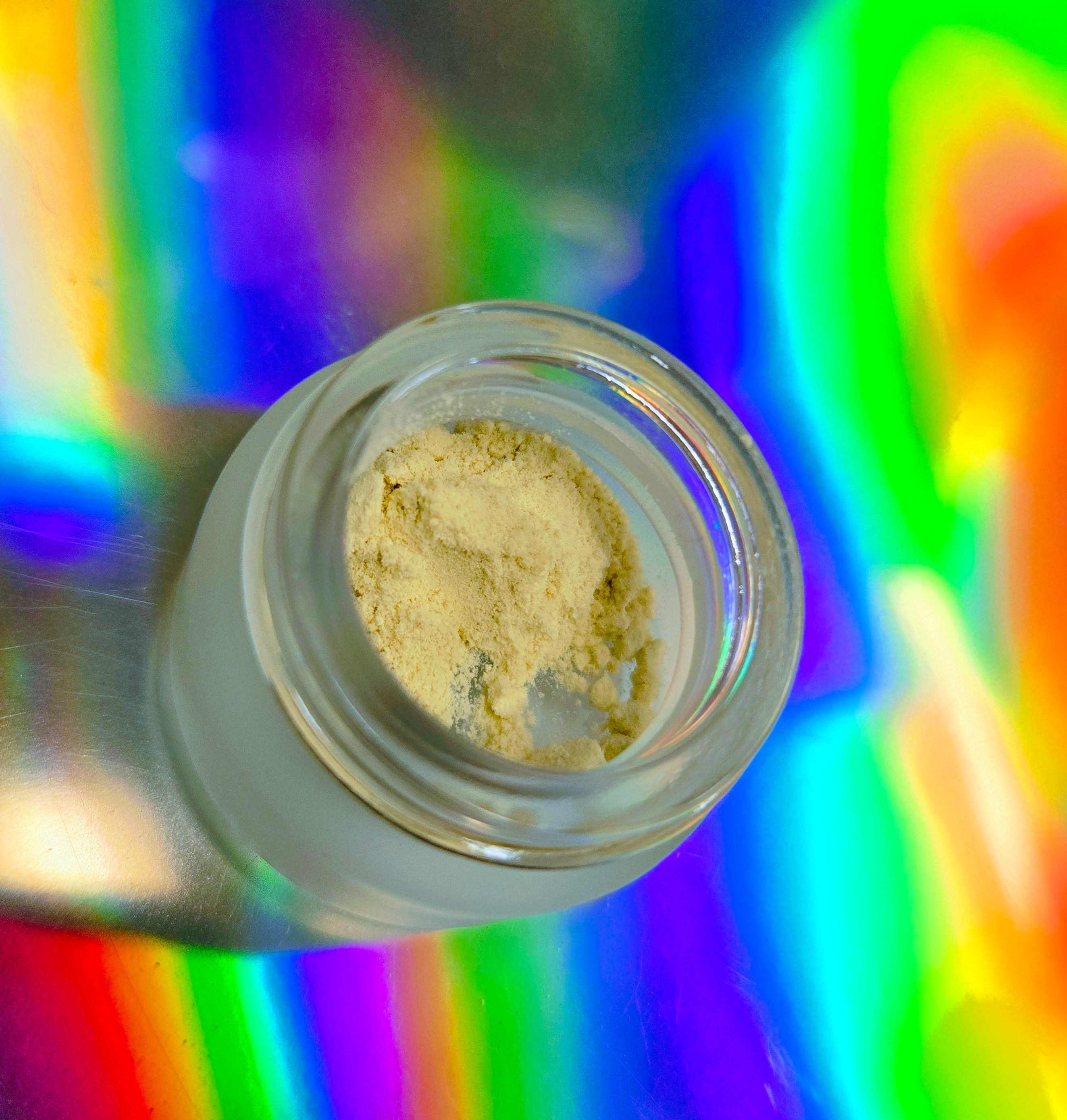 An open glass jar containing fine pale yellow kanna extract powder, set against a vivid holographic rainbow background.