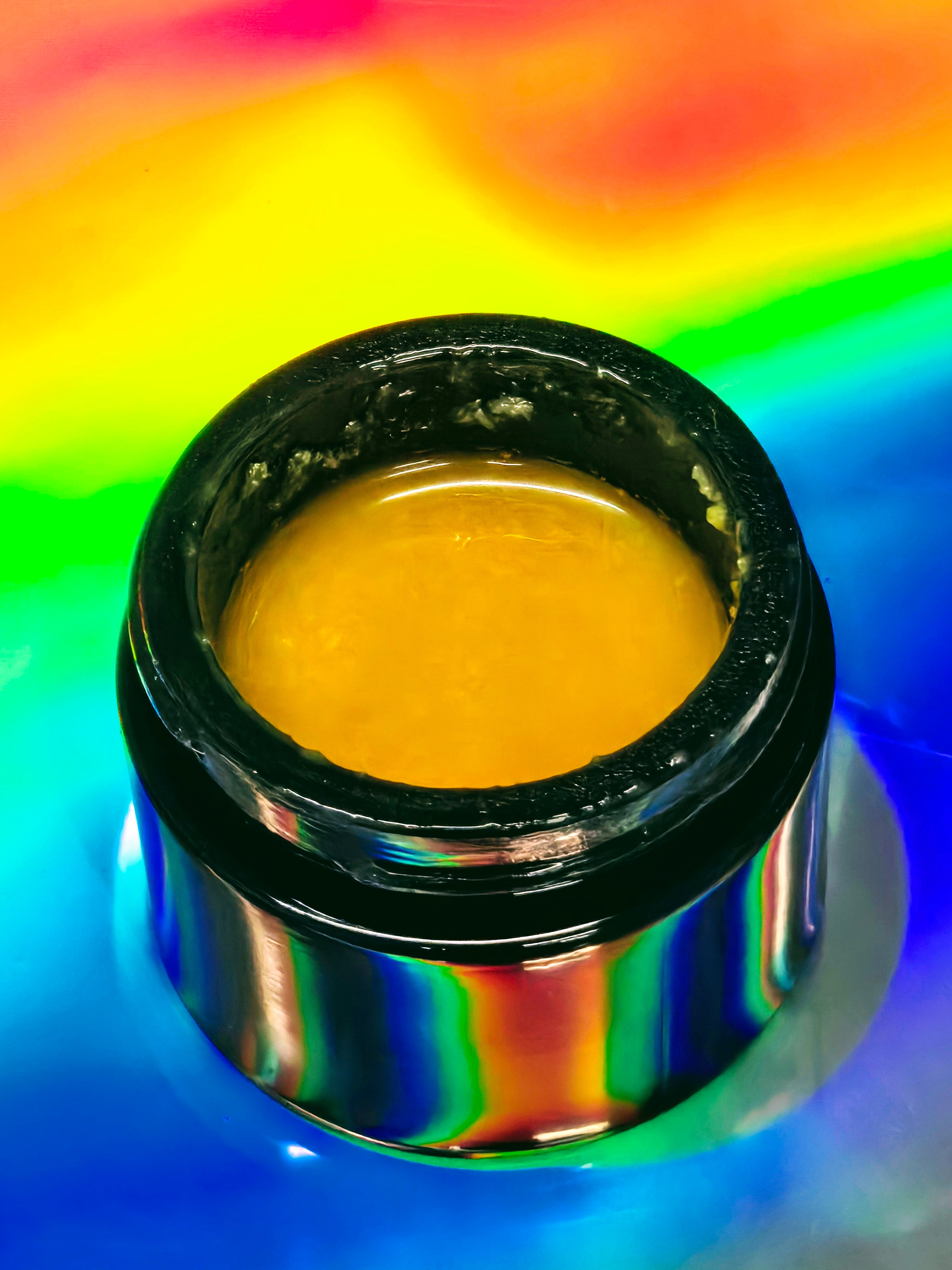 Small glass jar filled with smooth, golden-yellow kava CO₂ extract paste, shown with a glossy surface and thick texture, set against a vibrant holographic rainbow background that reflects colorful light around the container.