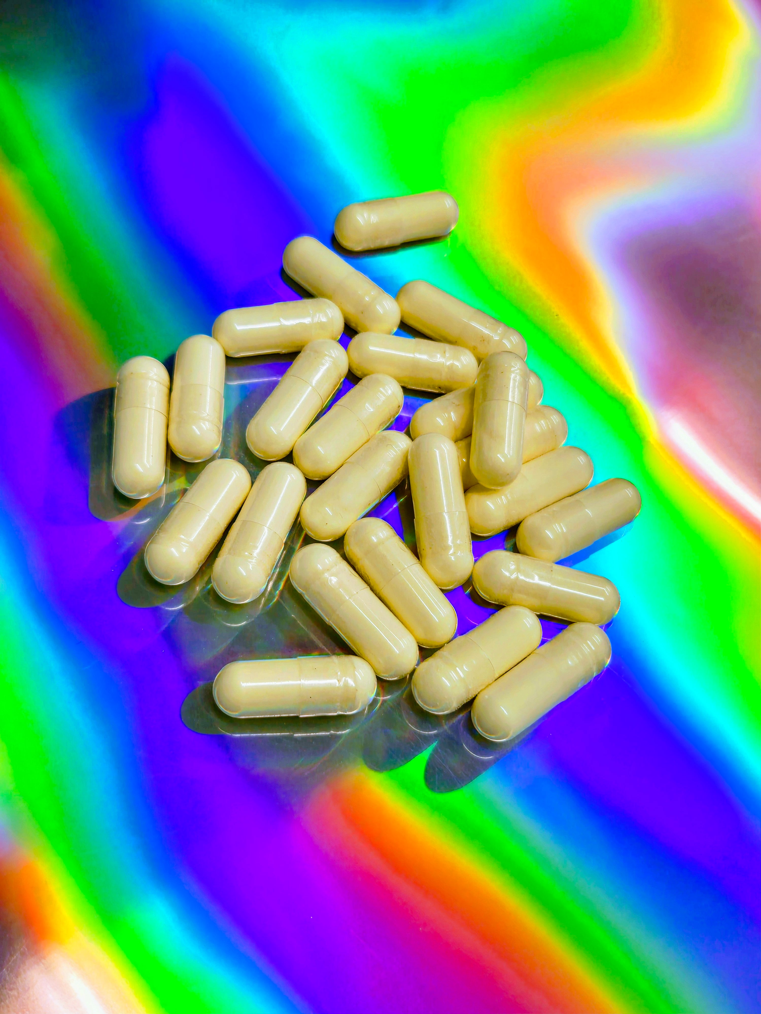 Scattered light-beige herbal capsules filled with powdered botanical extract, displayed on a vibrant rainbow holographic background.