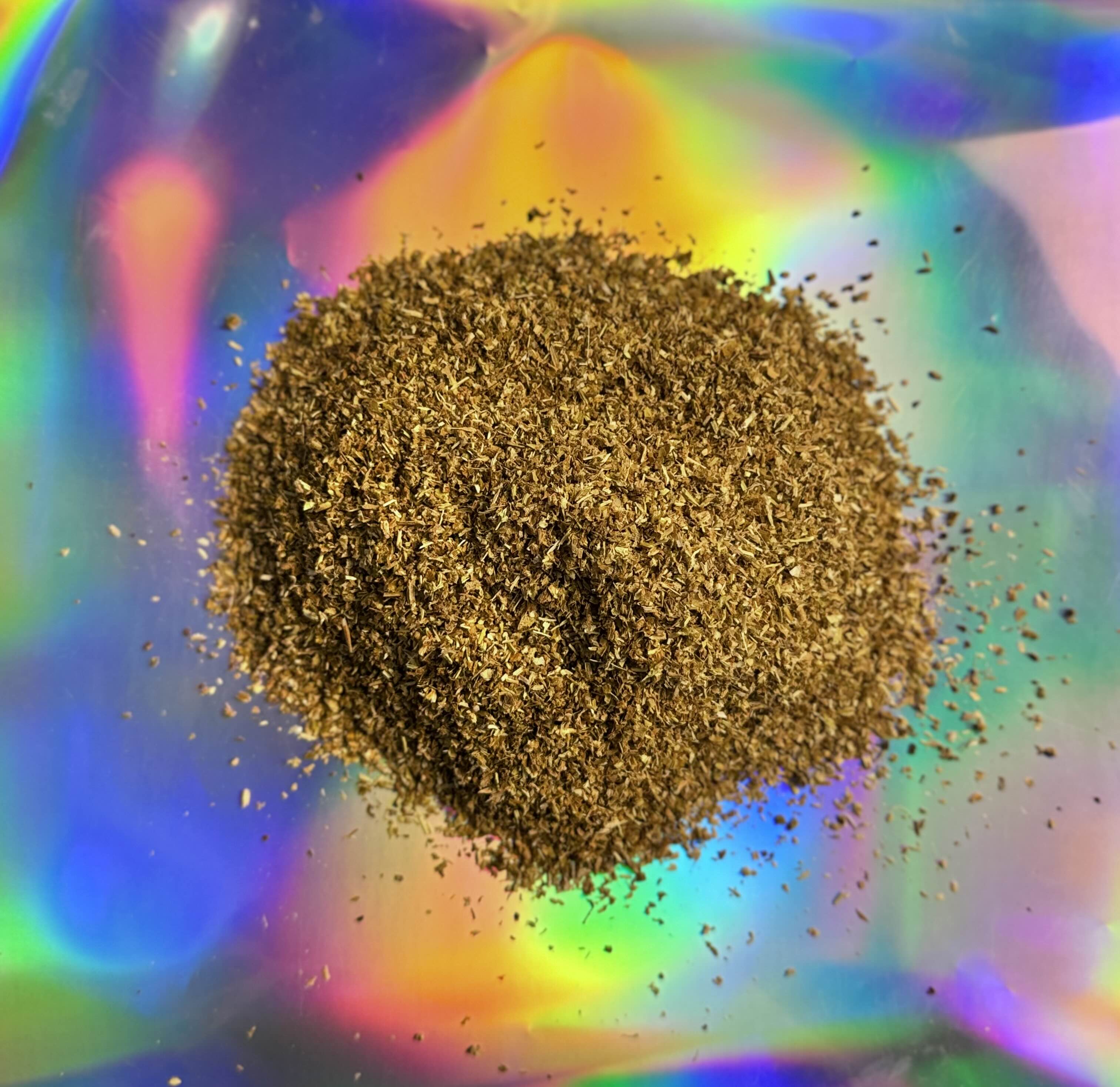 Close view of finely milled kanna leaf powder forming a loose mound with small particles suspended around it, set against a vivid holographic rainbow background.