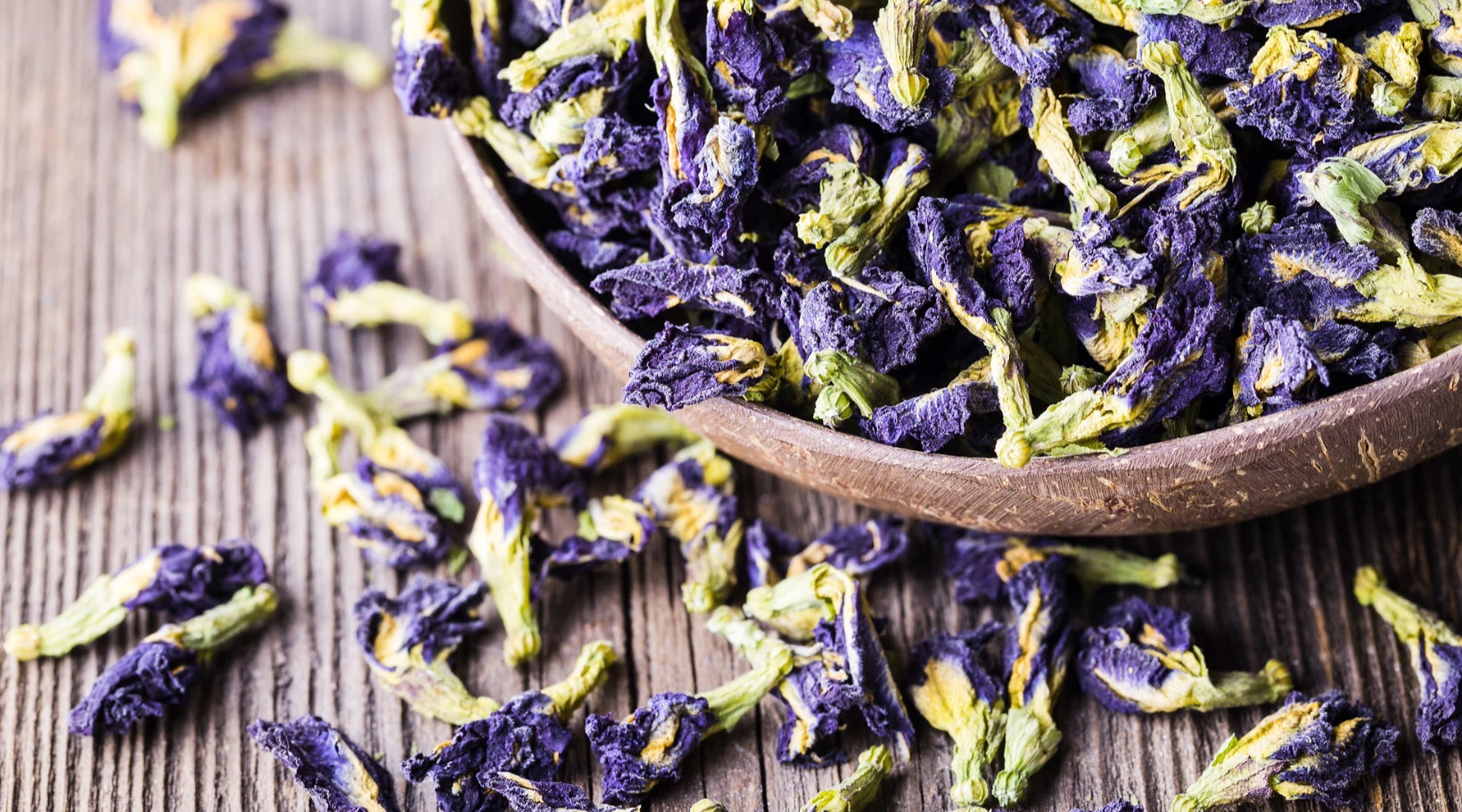 Organic butterfly pea flowers by Healing Herbals, showing vibrant dried blue and green blossoms in a wooden bowl with scattered petals on a rustic wooden surface.