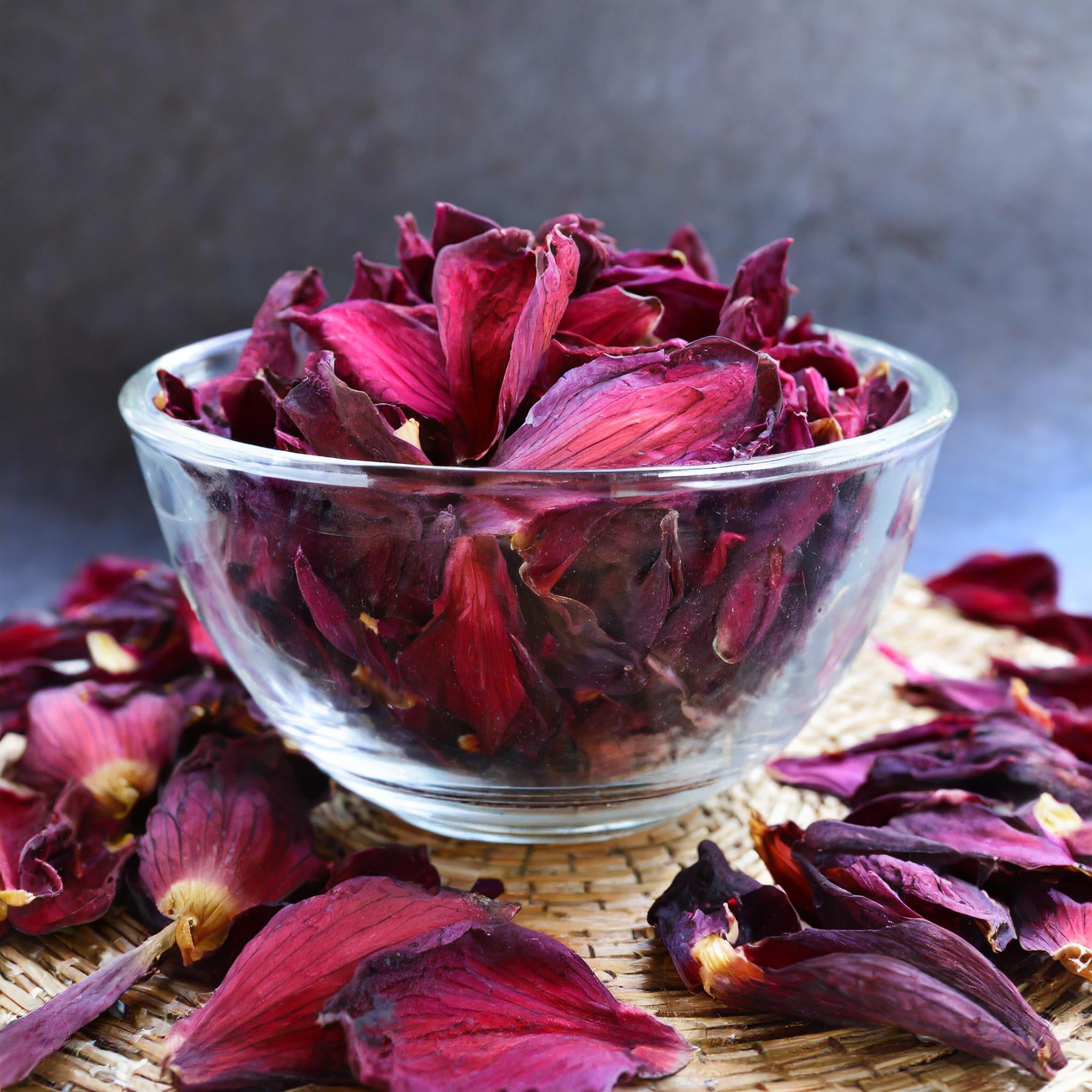 Red lotus loose-leaf tea by Healing Herbals, featuring deep crimson dried lotus petals in a clear glass bowl with scattered petals on a woven natural mat against a dark backdrop.