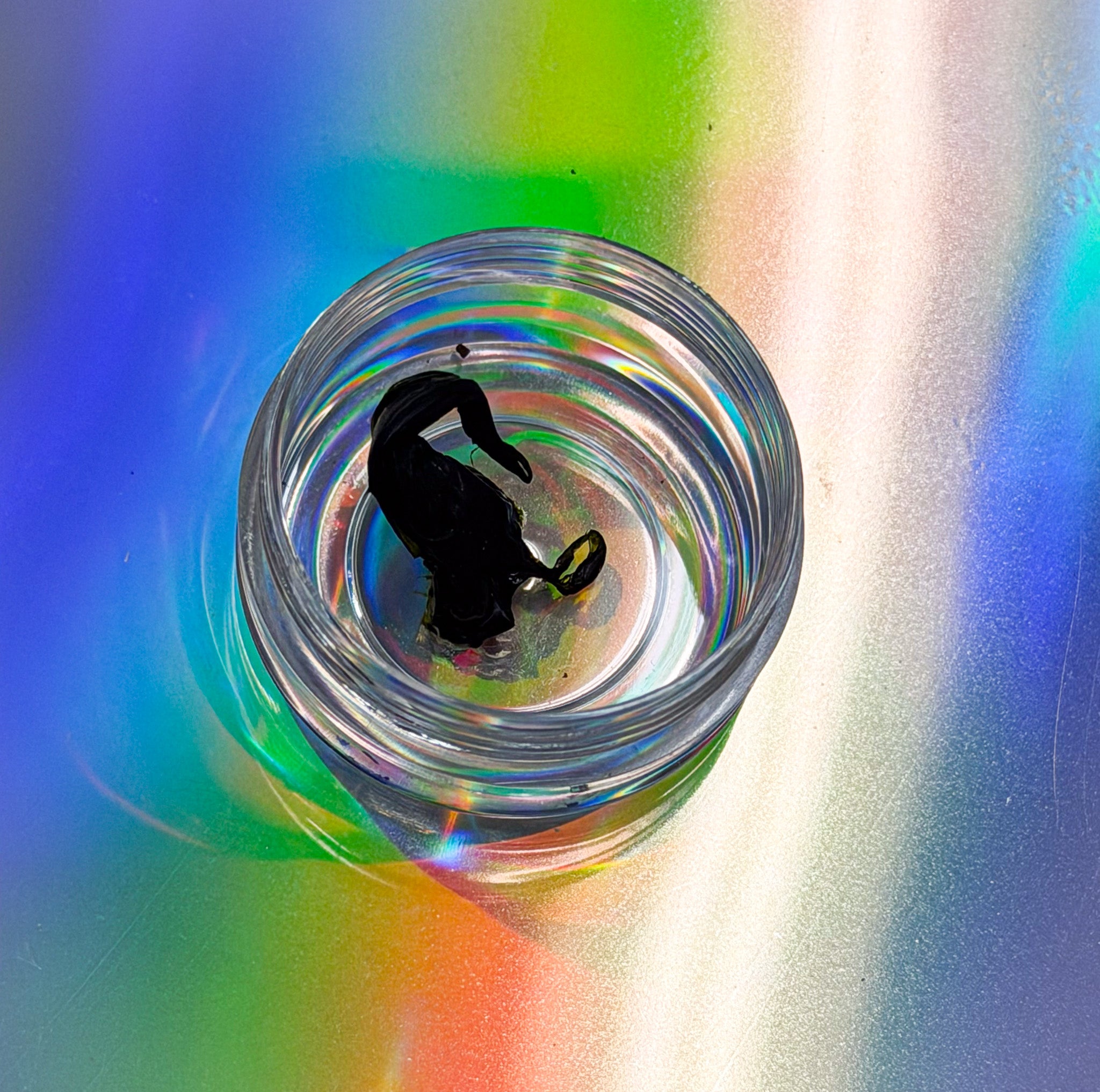 Pictured is a clear reflective jar with a dark stringy resin over a colorful pastel gradient. 