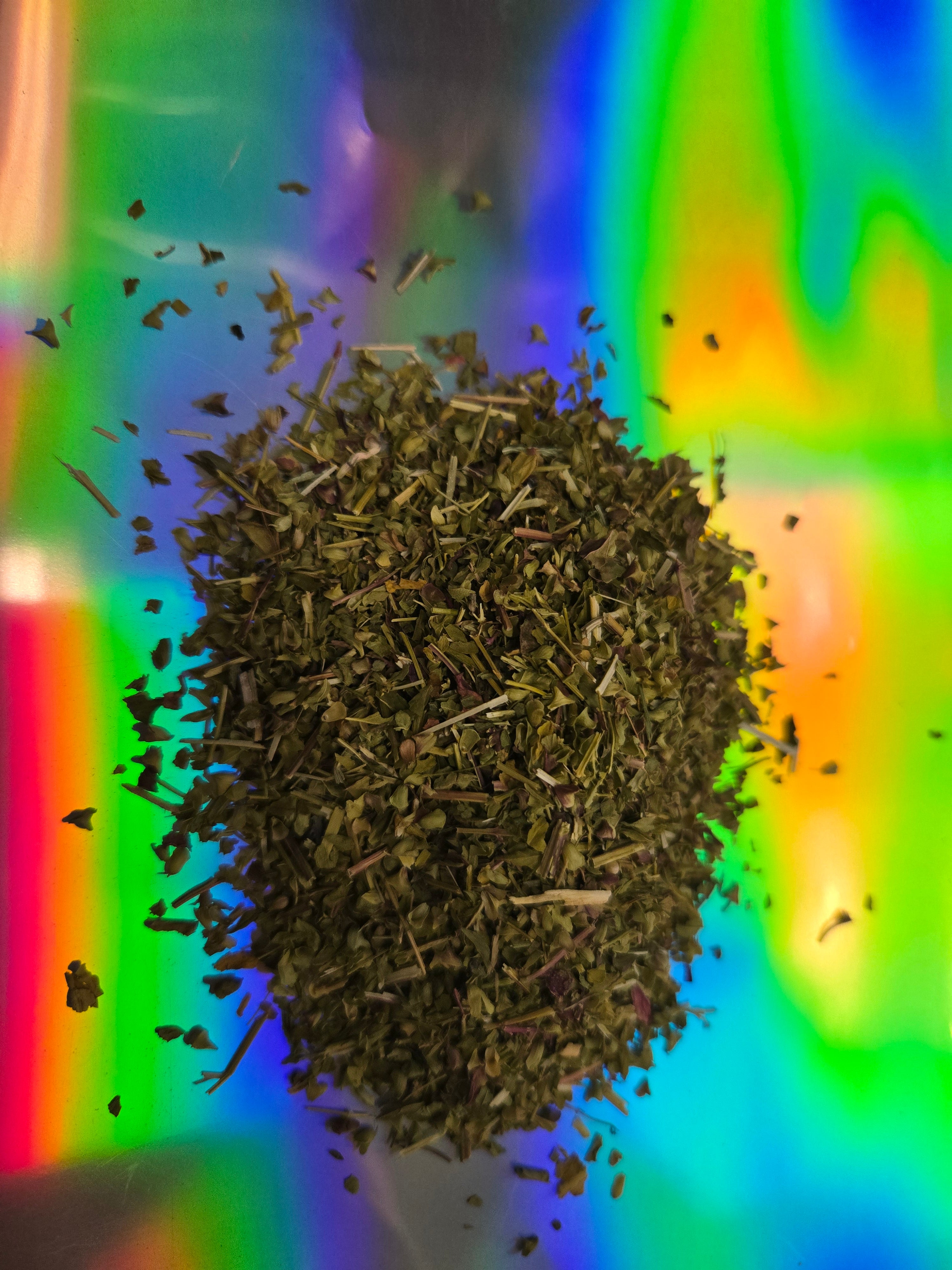 Top-down view of loose-leaf skullcap herb tea with finely cut green leaves and stems scattered mid-air over a vivid holographic rainbow background.