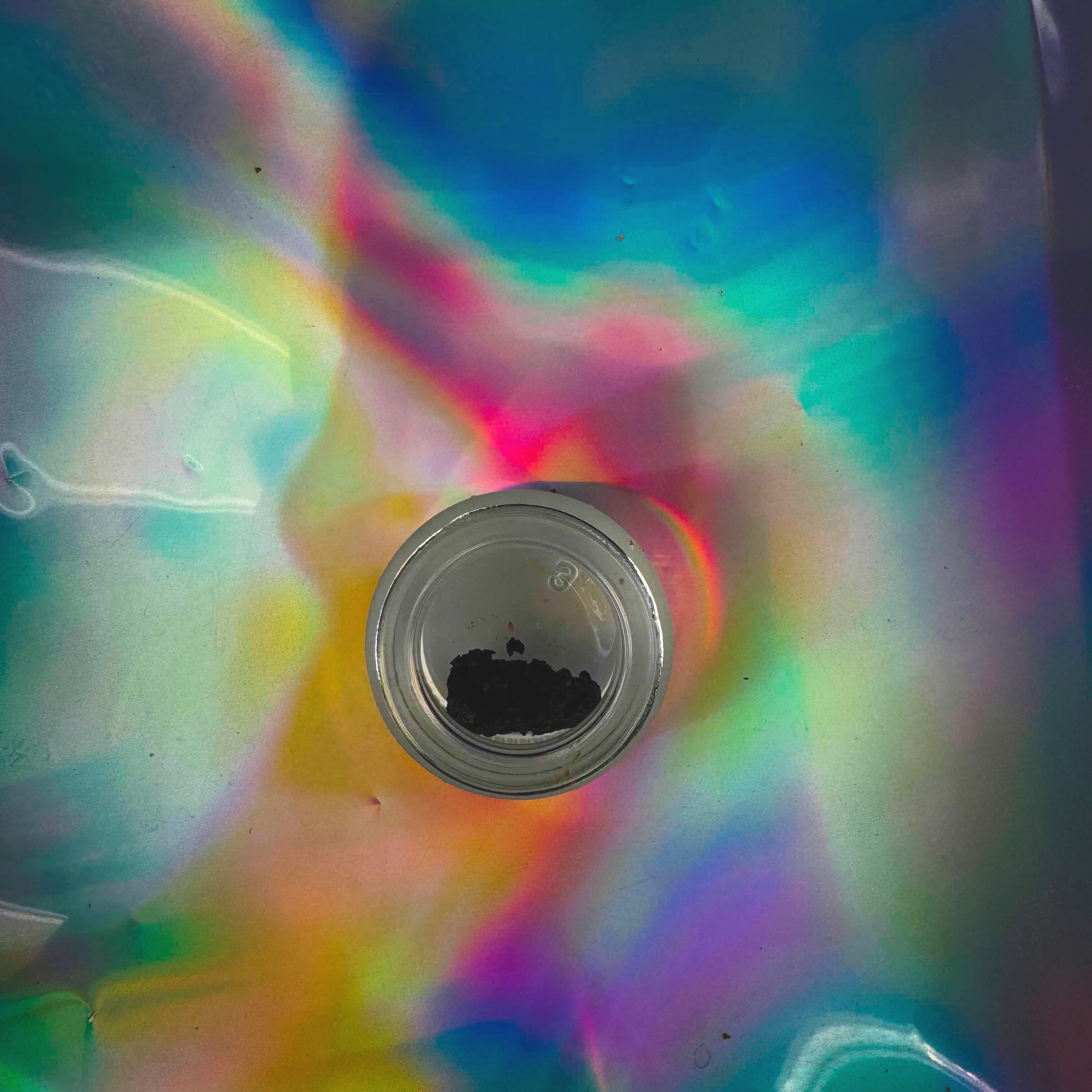 Top-down view of a small clear glass jar containing dark herbal resin extract on an iridescent holographic background.