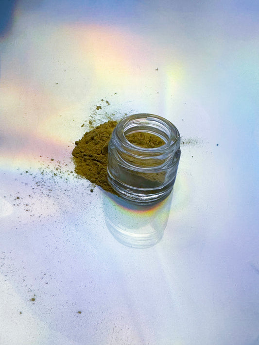 Small clear glass jar containing fine brown botanical powder, with a small pile spilled beside it on a light surface featuring soft rainbow reflections.