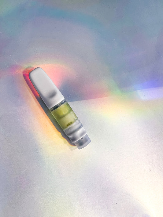 White vape cartridge filled with light green liquid, placed on a holographic rainbow light background.