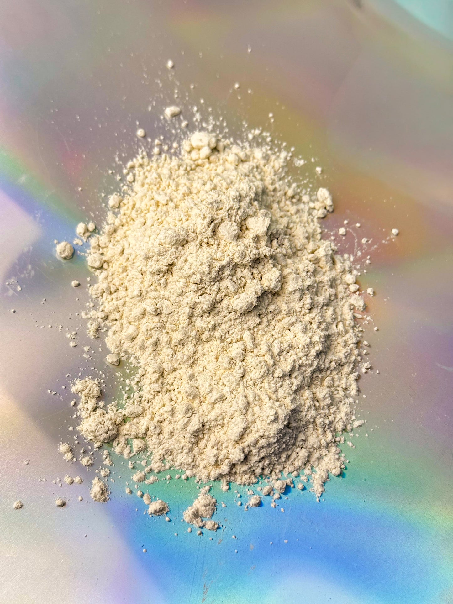Close-up of a small mound of fine off-white powder with soft clumps and scattered granules, displayed on a light surface with subtle rainbow reflections.