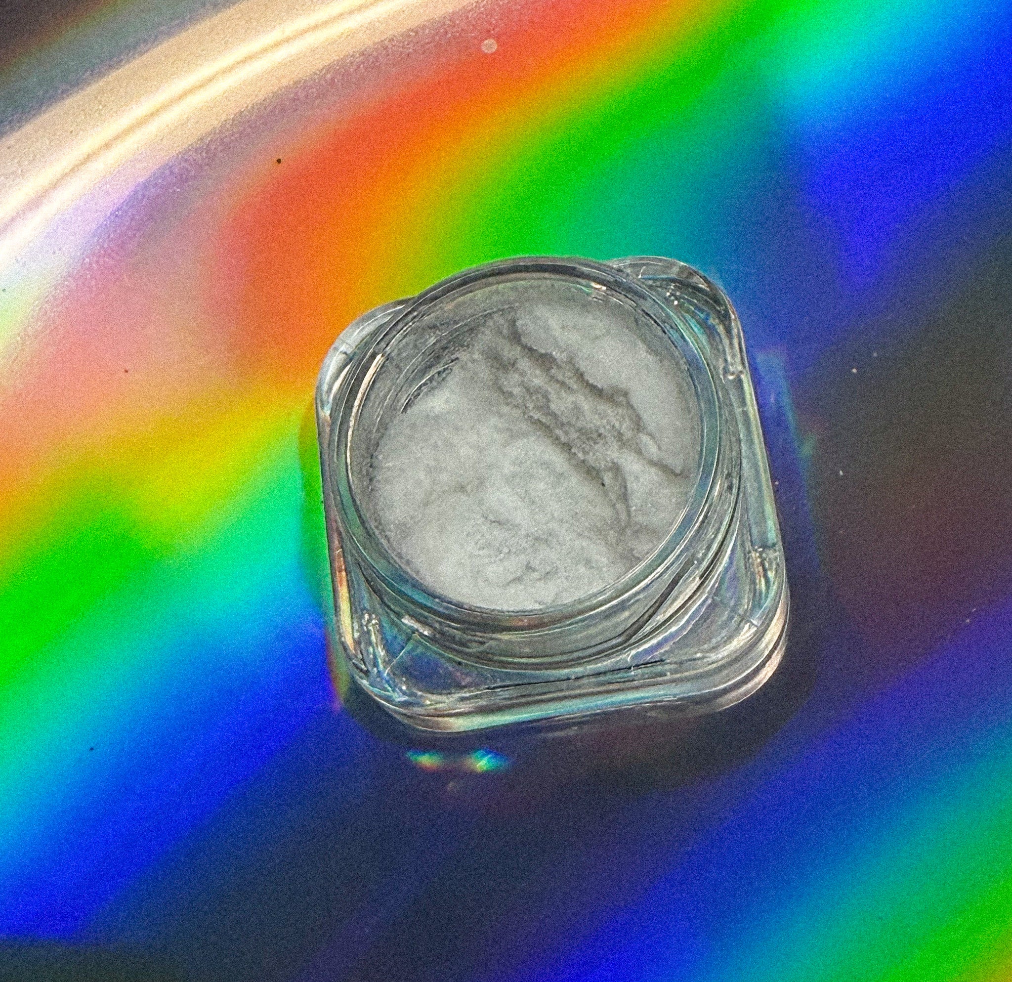 Small clear glass jar with grayish-white powder on a colorful, reflective surface.