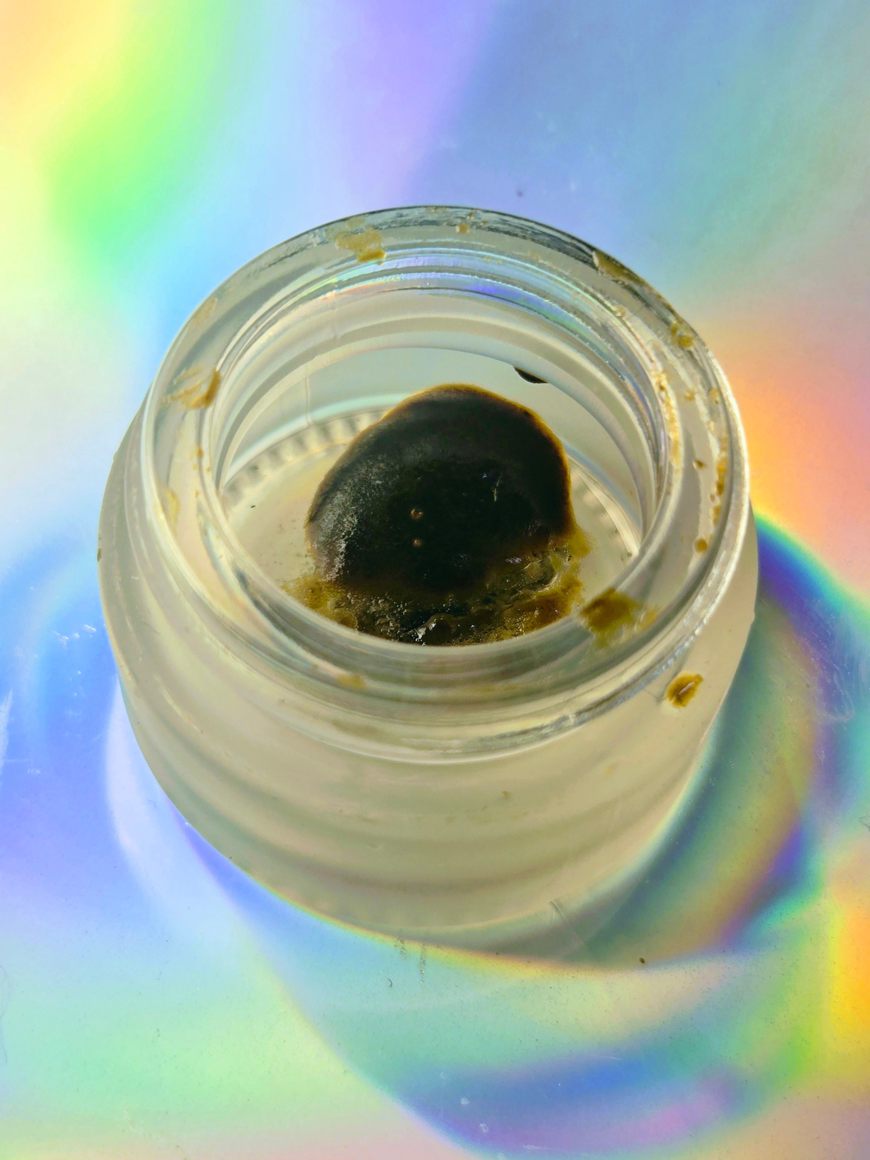 a small clear glass jar holding dark, shiny herbal resin extract, placed on a holographic surface with rainbow light reflections.
