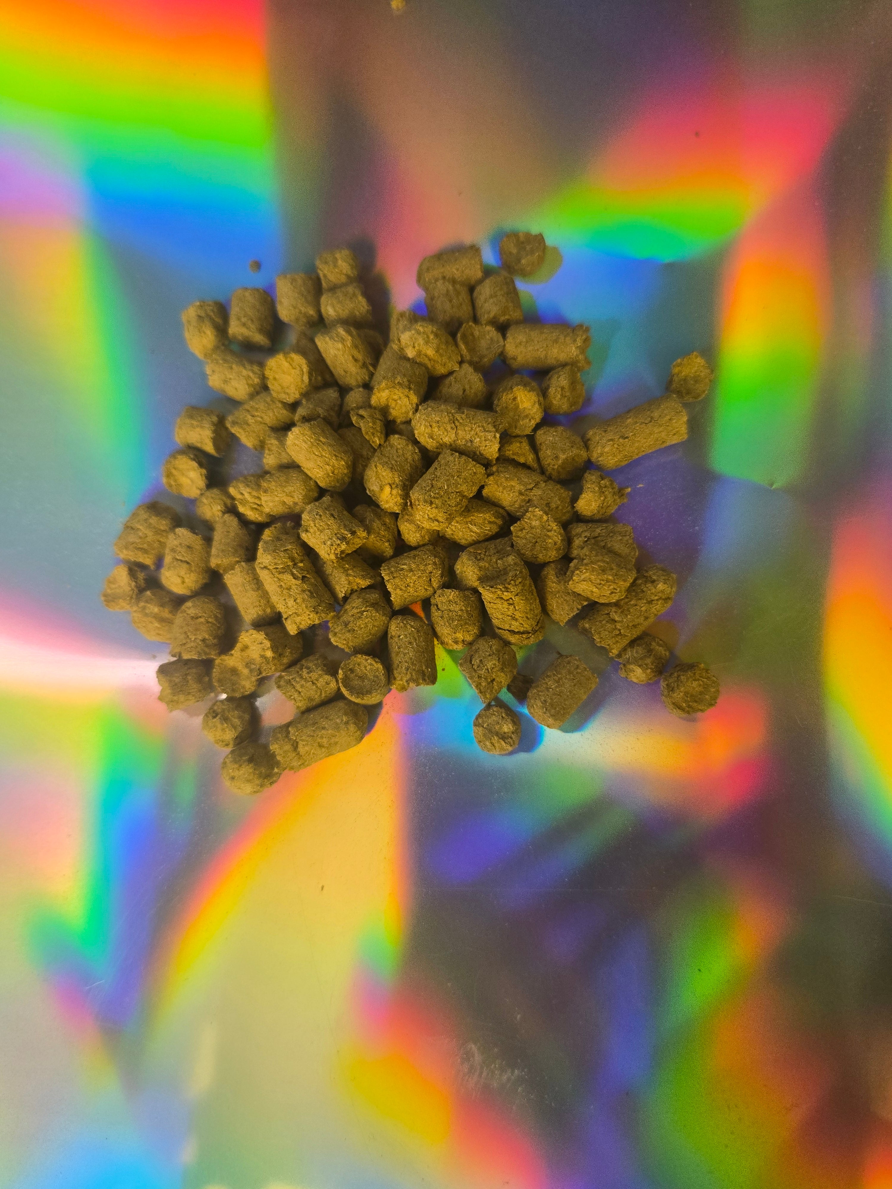 Close-up of small cylindrical herbal pellets clustered together, showing a compact, earthy texture, displayed on a reflective holographic background with rainbow light patterns.