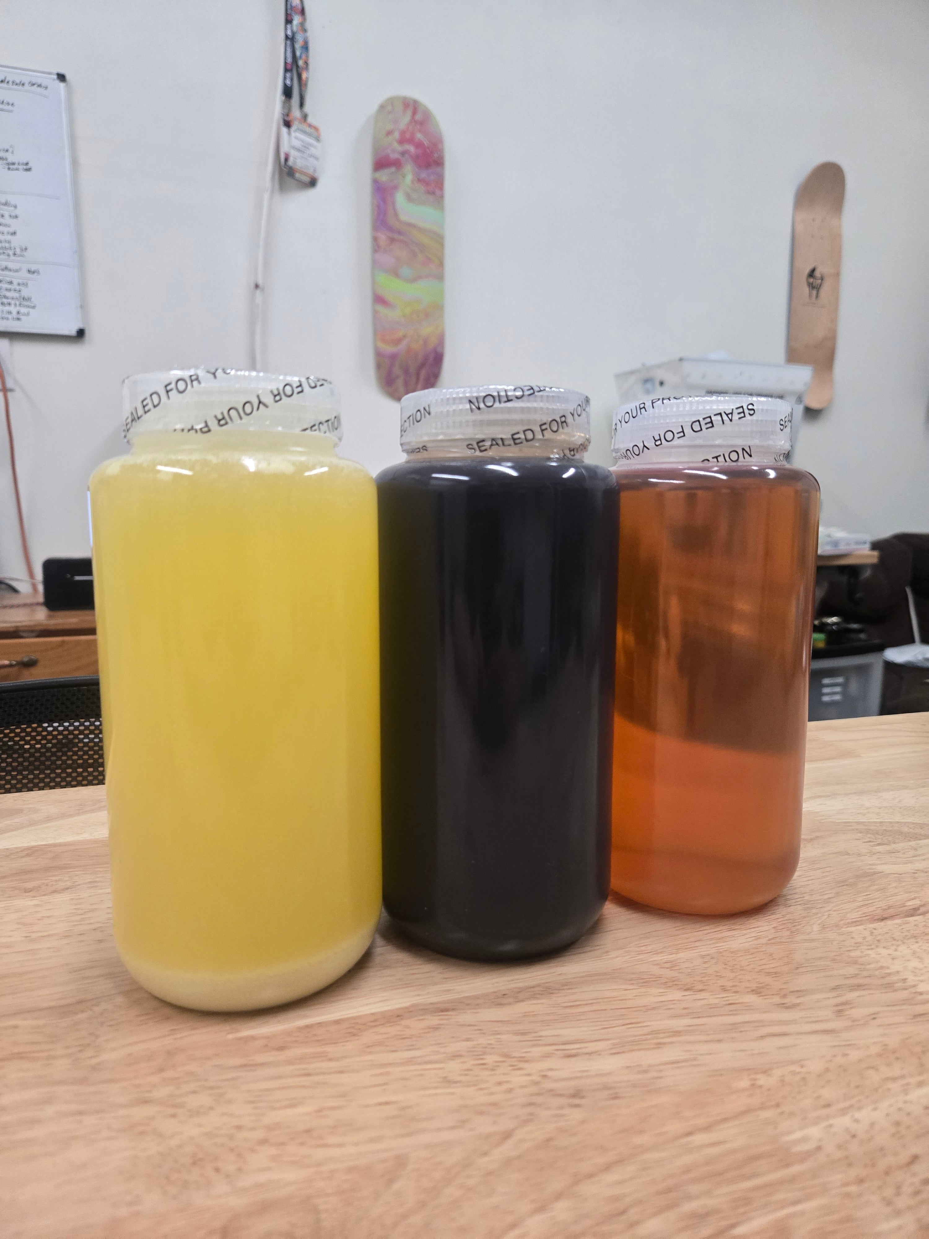 Three sealed glass jars filled with liquids pale yellow, dark black, and amber arranged side by side on a wooden table, with a workshop interior and skateboard decks visible in the background.