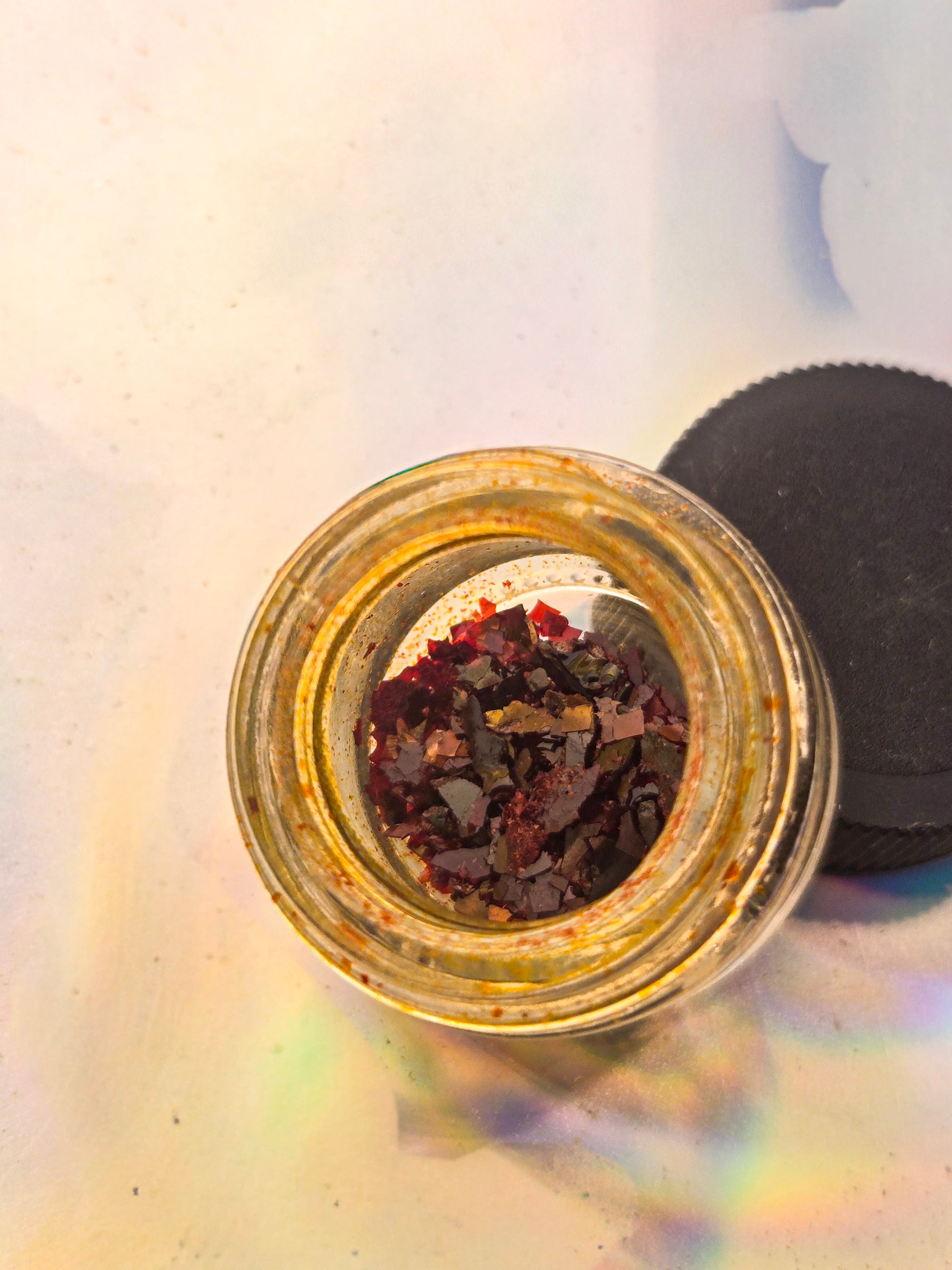 Top view of a small glass jar filled with dark red resin flakes, placed on a holographic surface with lid beside it.