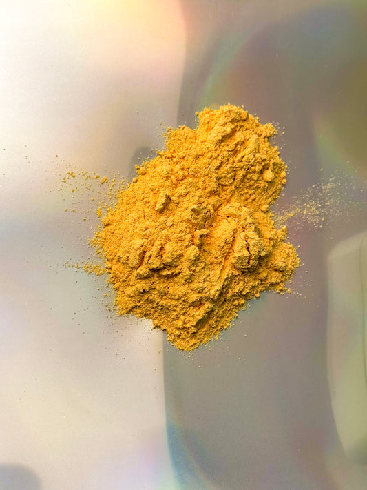 Close-up of a loose pile of fine golden-yellow botanical powder with small natural clumps, displayed on a light surface with soft rainbow reflections.